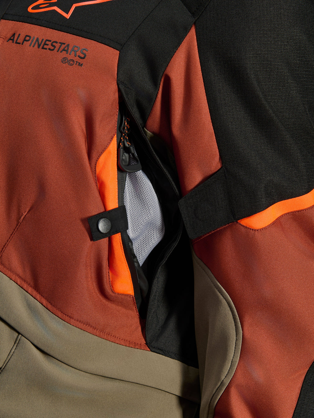 Veste Alpinestars Andes V4 Drystar®, Walnut Rust Black, gros plan sur l'ouverture de ventilation directe avec doublure en maille, textile durable avec accents orange, bouton-pression et détail du logo Alpinestars