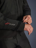 Veste Alpinestars Andes V4 Drystar®, veste de moto en textile noir, gros plan sur la manche arborant les logos Alpinestars rouge et Drystar® blanc, pilote ajustant la sangle velcro au poignet pour un maintien sûr, détail fonctionnel pour la conduite aventure