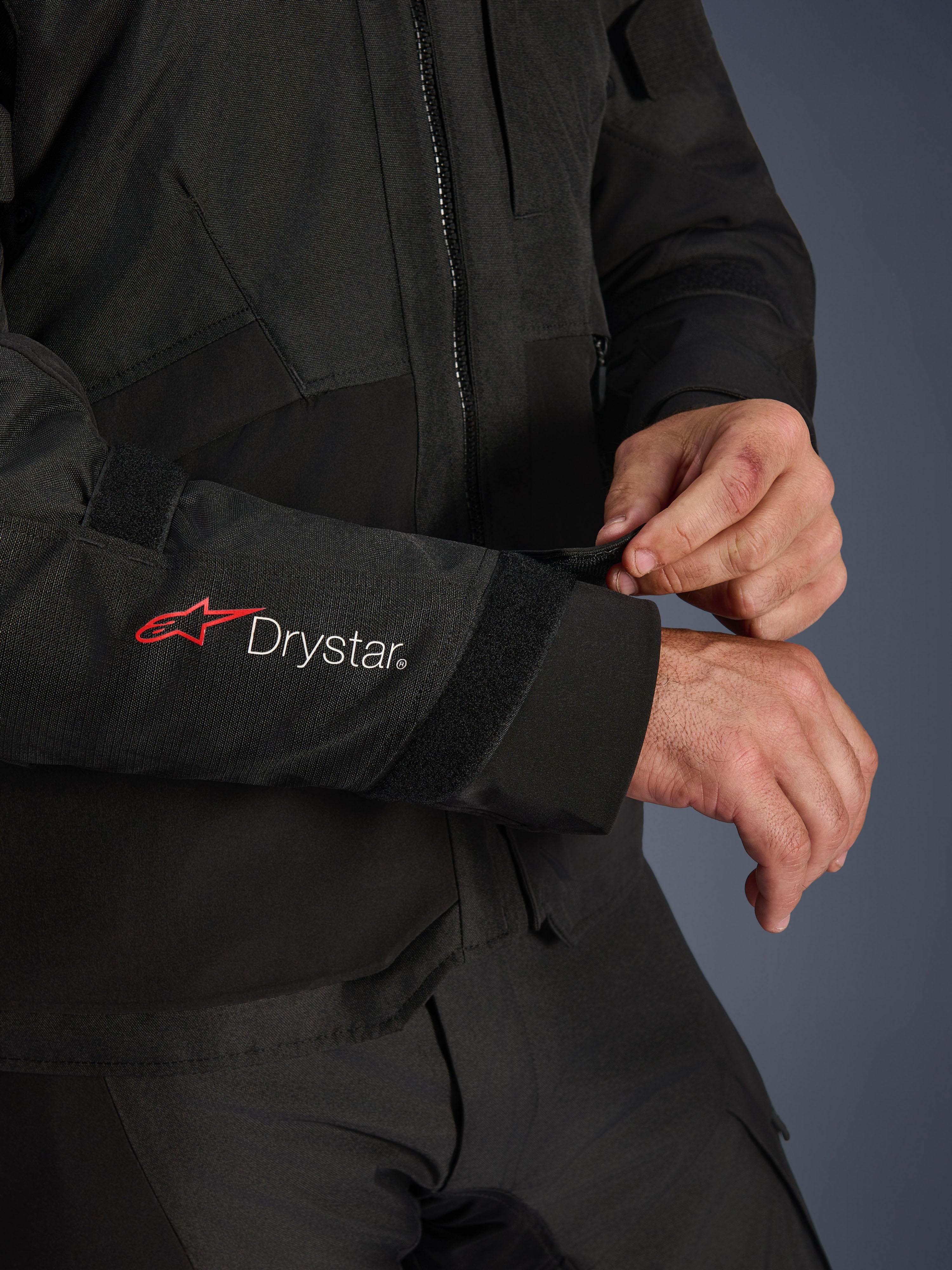 Veste Alpinestars Andes V4 Drystar®, veste de moto en textile noir, gros plan sur la manche arborant les logos Alpinestars rouge et Drystar® blanc, pilote ajustant la sangle velcro au poignet pour un maintien sûr, détail fonctionnel pour la conduite aventure