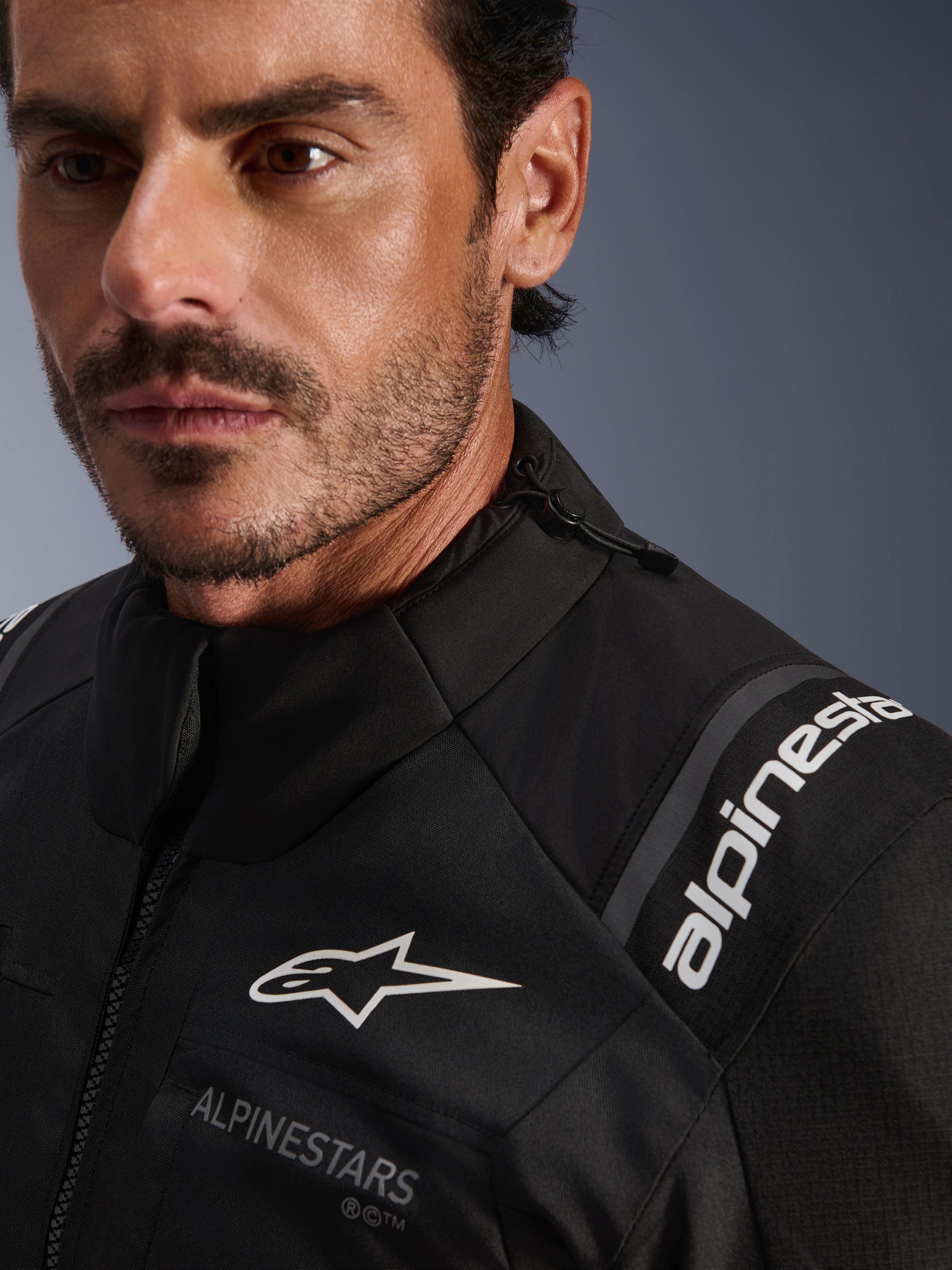 Veste Alpinestars Andes V4 Drystar®, veste de moto en textile noir, gros plan détaillé porté par un homme, avec logos Alpinestars blancs sur la poitrine et l'épaule, cordon de serrage réglable au col, conception adventure-touring haute performance.