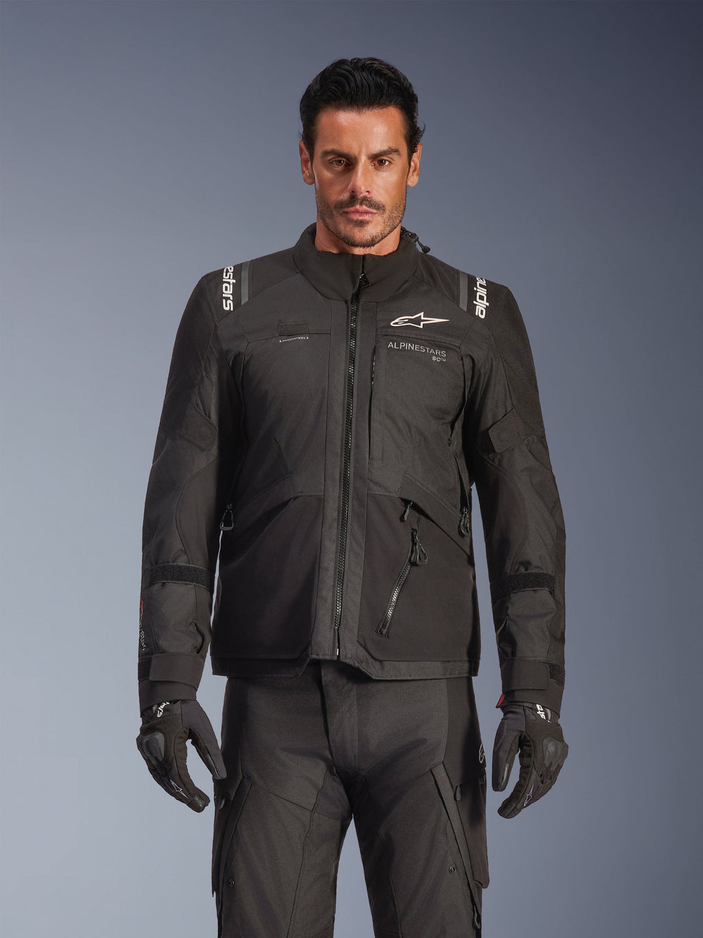 Mannequin masculin portant la veste Alpinestars Andes V4 Drystar® en noir, veste de moto d'aventure touring, avec pantalon et gants de moto noirs assortis, fermetures éclair imperméables et logo Alpinestars blanc sur la poitrine et les épaules, équipement de protection toutes saisons, prise de vue en studio