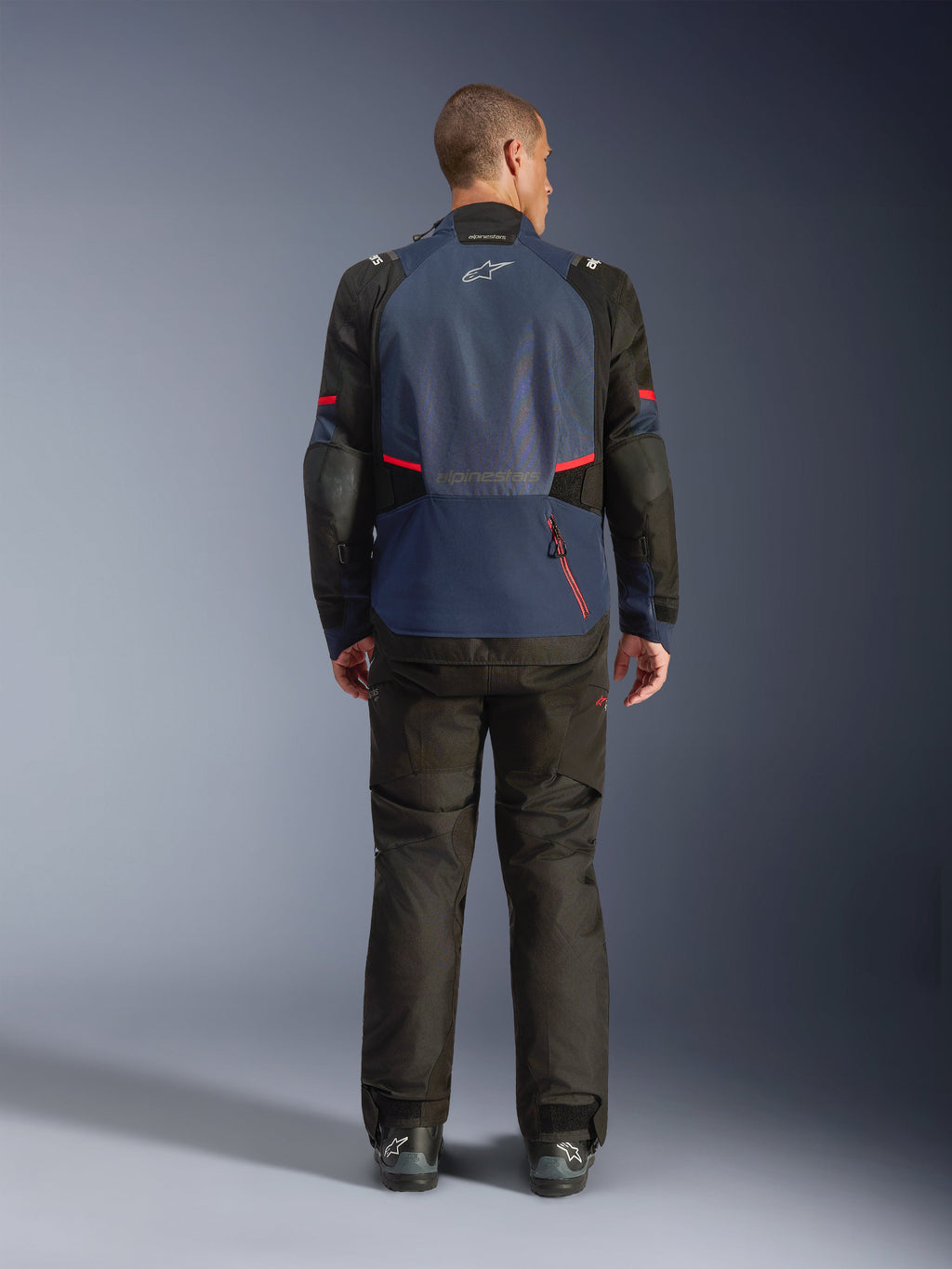 Veste Alpinestars Andes V4 Drystar®, vue de dos, bleu Night Navy et Mood Indigo avec panneaux noirs et accents rouges, portée avec un pantalon noir assorti, avec le logo Alpinestars sur le haut du dos et une grande fermeture éclair pour le compartiment arrière.