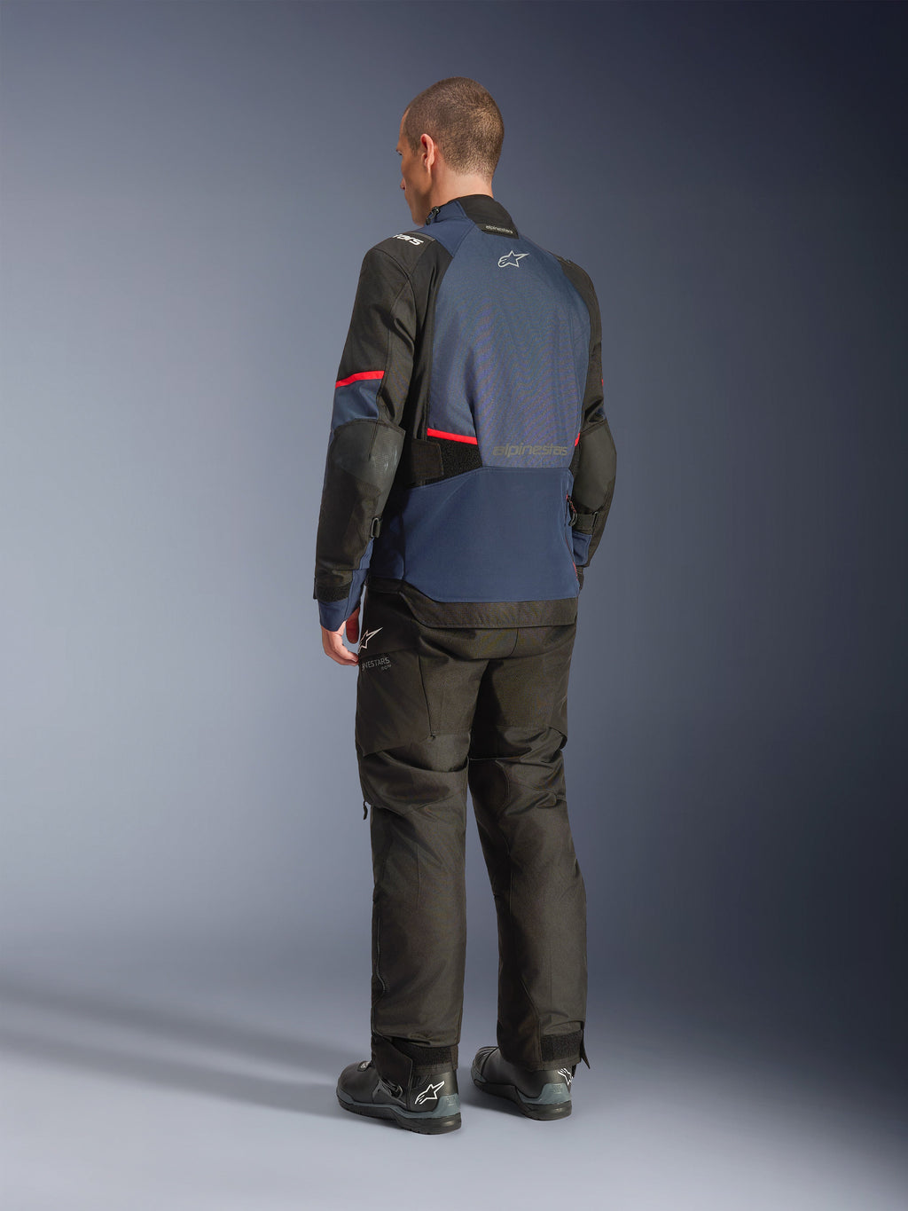 Veste Alpinestars Andes V4 Drystar®, Night Navy et Mood Indigo avec empiècements noirs et accents rouges, vue de dos de trois-quarts, portée avec un pantalon de moto d'aventure et des bottes noirs assortis, construction en textile technique avec logo Alpinestars sur le haut du dos, ourlet arrière allongé pour une meilleure protection.