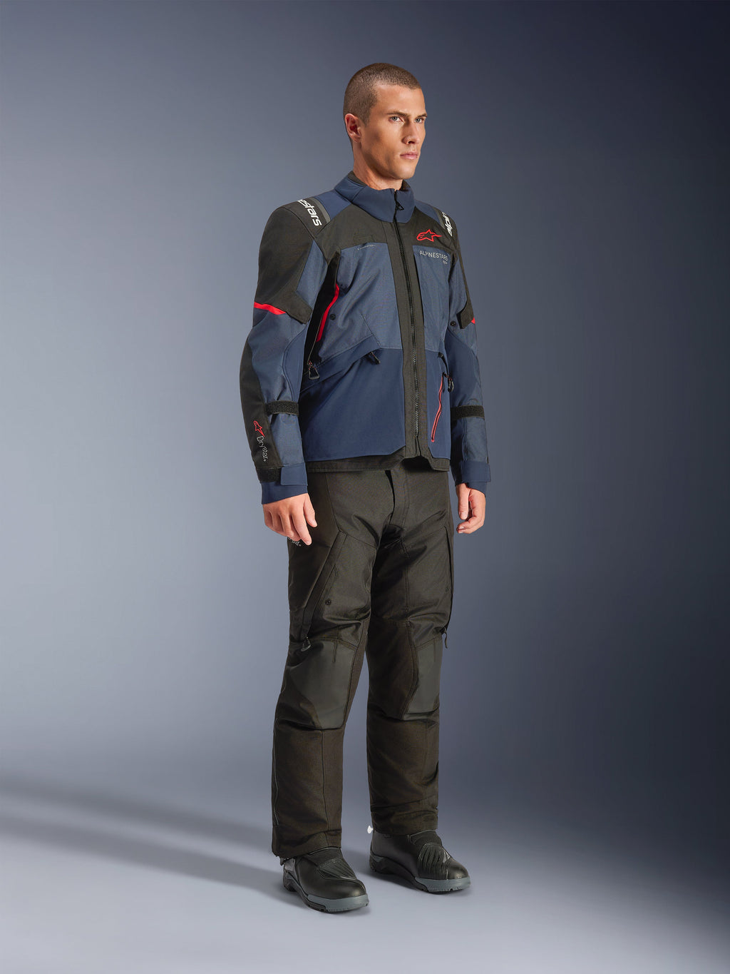 Modèle masculin portant la veste Alpinestars Andes V4 Drystar® en Night Navy et Mood Indigo avec empiècements noirs et accents rouges, associée à un pantalon de moto et des bottes noirs, équipement technique de touring d'aventure en studio.