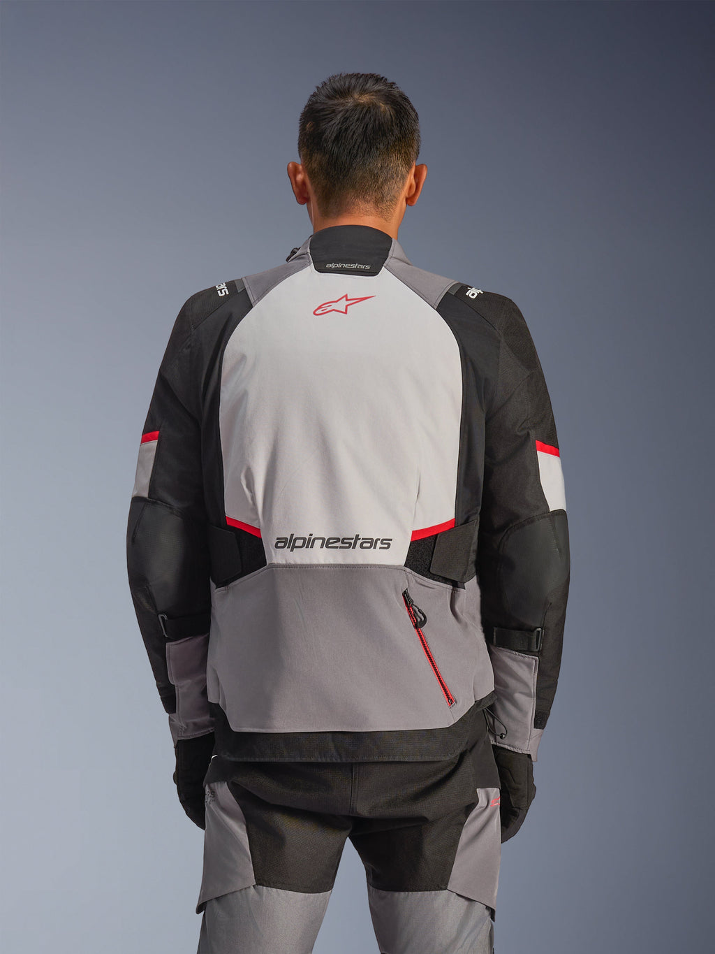 Veste Alpinestars Andes V4 Drystar®, vue de dos, gris foncé, gris glace, noir et rouge vif, arborant un logo Astar rouge et le lettrage Alpinestars dans le dos, avec des panneaux renforcés et une poche zippée rouge, équipement de moto d'aventure