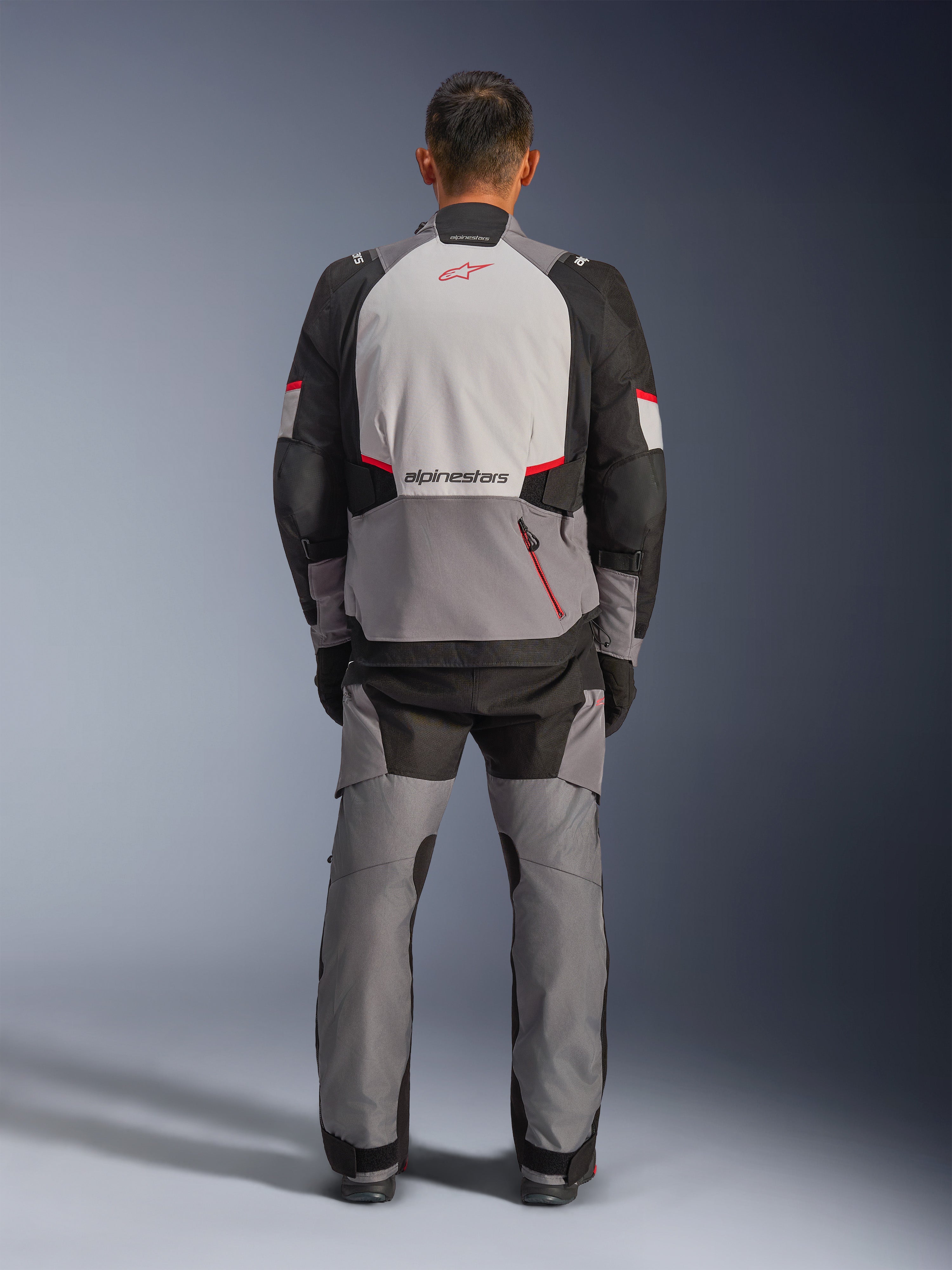 Veste Alpinestars Andes V4 Drystar®, veste de moto d'aventure, gris foncé, gris glacé, noir et rouge vif, vue arrière d'un pilote portant un pantalon assorti, avec un large panneau dorsal gris glacé et logo rouge, sections d'épaules renforcées noires et sangles de réglage à la taille pour le touring toutes saisons.