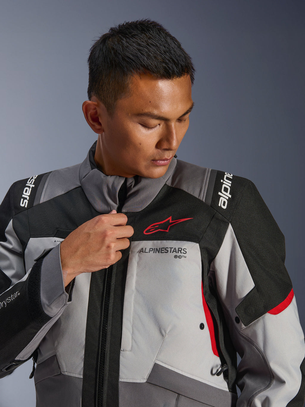 Mannequin portant la veste Alpinestars Andes V4 Drystar®, gris foncé et gris glace avec des accents noirs et rouge vif, ajustant la fermeture éclair frontale, arborant le logo Astars rouge et le marquage Drystar®, style touring aventure