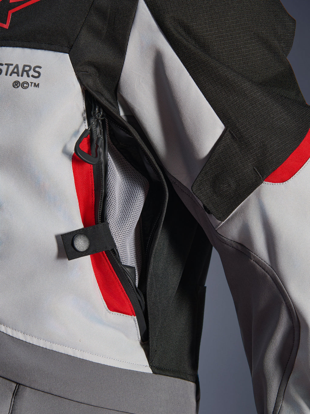 Gros plan sur les détails de la veste Alpinestars Andes V4 Drystar® en gris foncé, gris glacier et rouge vif, mettant en évidence une ouverture de ventilation zippée sur la poitrine avec doublure en mesh et une sangle de réglage à bouton-pression.