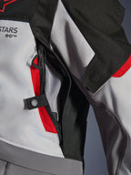 Gros plan sur les détails de la veste Alpinestars Andes V4 Drystar® en gris foncé, gris glacier et rouge vif, mettant en évidence une ouverture de ventilation zippée sur la poitrine avec doublure en mesh et une sangle de réglage à bouton-pression.