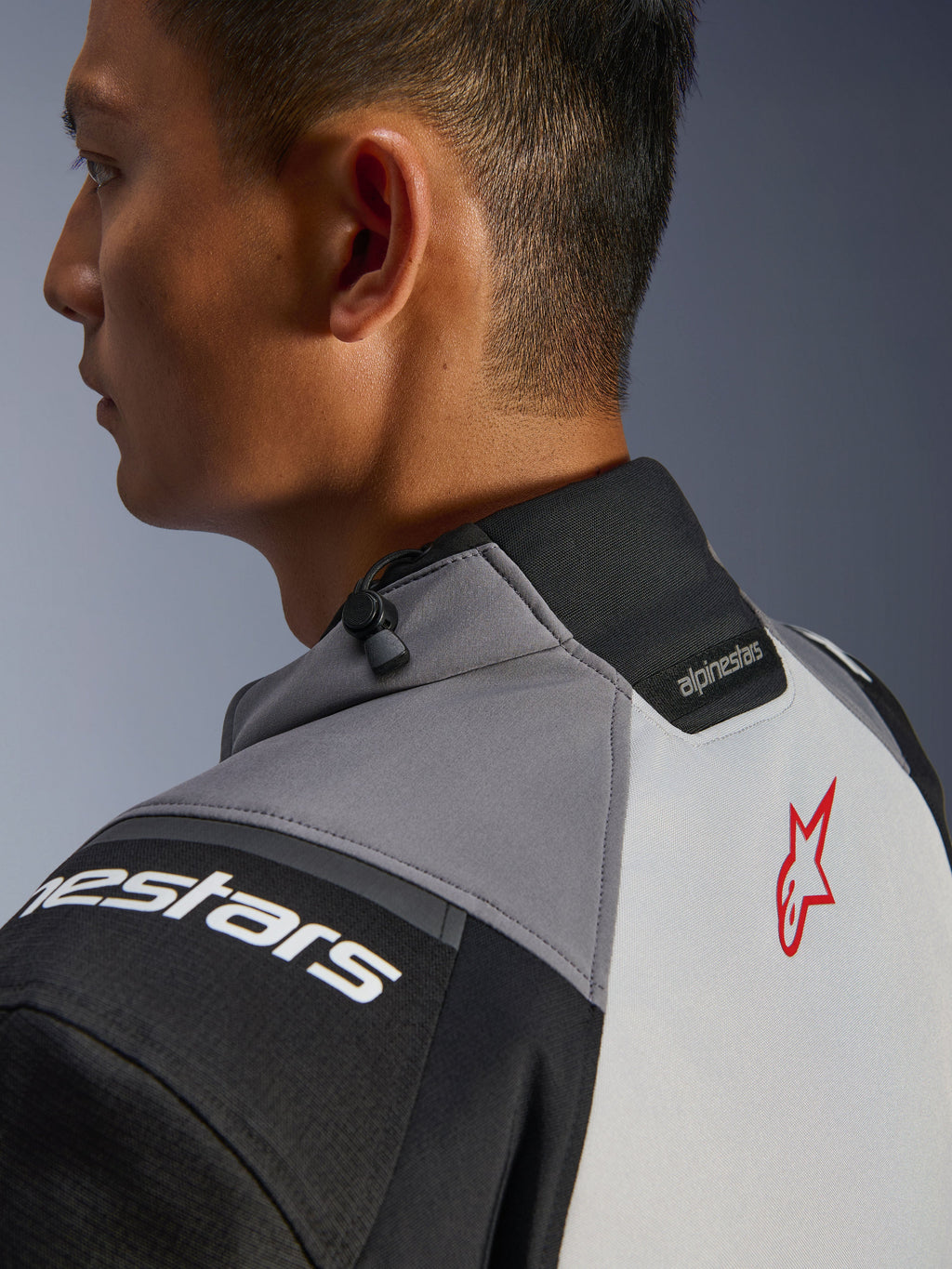 Veste Alpinestars Andes V4 Drystar®, gros plan sur le col et le haut du dos, gris foncé et gris glace avec détails noirs, logo étoile rouge, dotée d'un cordon de serrage au col et du marquage Alpinestars, construction textile durable