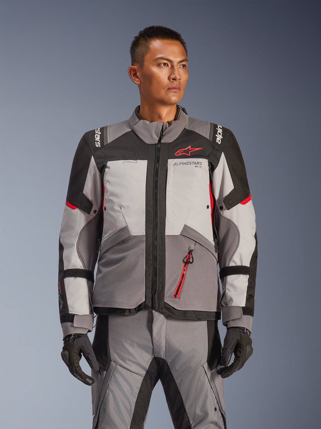Veste Alpinestars Andes V4 Drystar®, veste de moto touring aventure, gris foncé et gris glace avec des accents noirs et rouge vif, portée avec un pantalon et des gants assortis, conception imperméable et respirante pour la conduite d'aventure par tous les temps