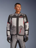 Veste Alpinestars Andes V4 Drystar®, veste de moto touring aventure, gris foncé et gris glace avec des accents noirs et rouge vif, portée avec un pantalon et des gants assortis, conception imperméable et respirante pour la conduite d'aventure par tous les temps