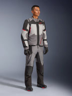 Modèle masculin portant la veste Alpinestars Andes V4 Drystar® et le pantalon d'aventure assorti, coloris gris foncé, gris glace, noir et rouge vif, avec des gants et des bottes de moto noirs, équipement technique toutes saisons au design multi-panneaux moderne.