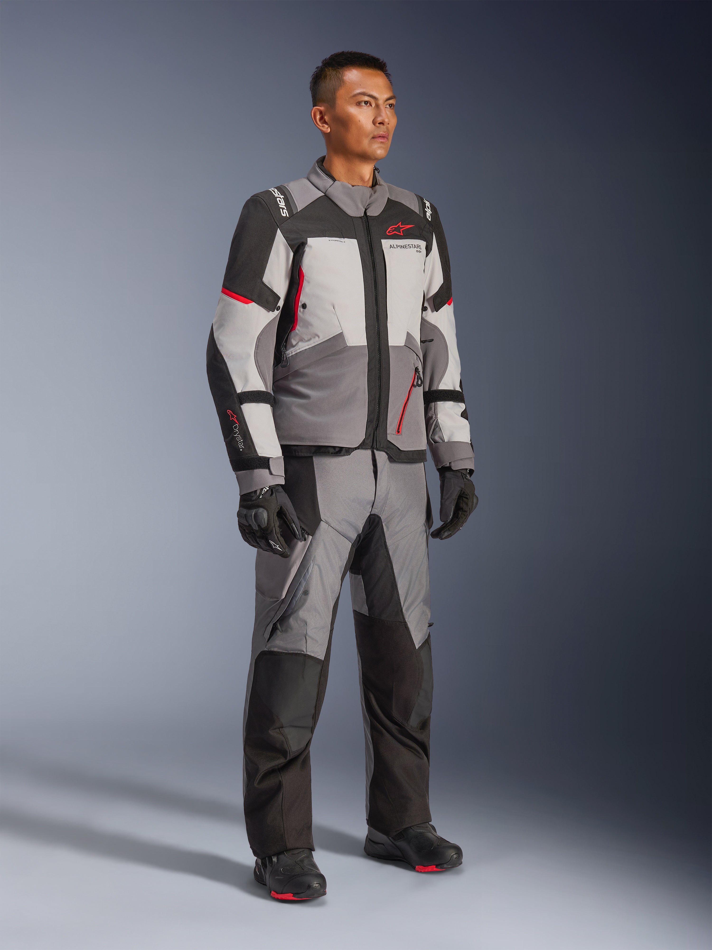 Modèle masculin portant la veste Alpinestars Andes V4 Drystar® et le pantalon d'aventure assorti, coloris gris foncé, gris glace, noir et rouge vif, avec des gants et des bottes de moto noirs, équipement technique toutes saisons au design multi-panneaux moderne.