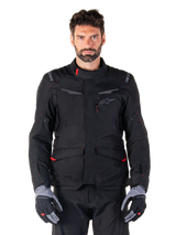 Veste imperméable Alpinestars ST-1, veste de touring d'aventure moto noire, portée par un mannequin homme avec un pantalon noir assorti et des gants gris/noir, style minimaliste, logo Alpinestars sur les épaules, grandes poches cargo avec détails rouges, conception ergonomique pour le touring routier par tous les temps
