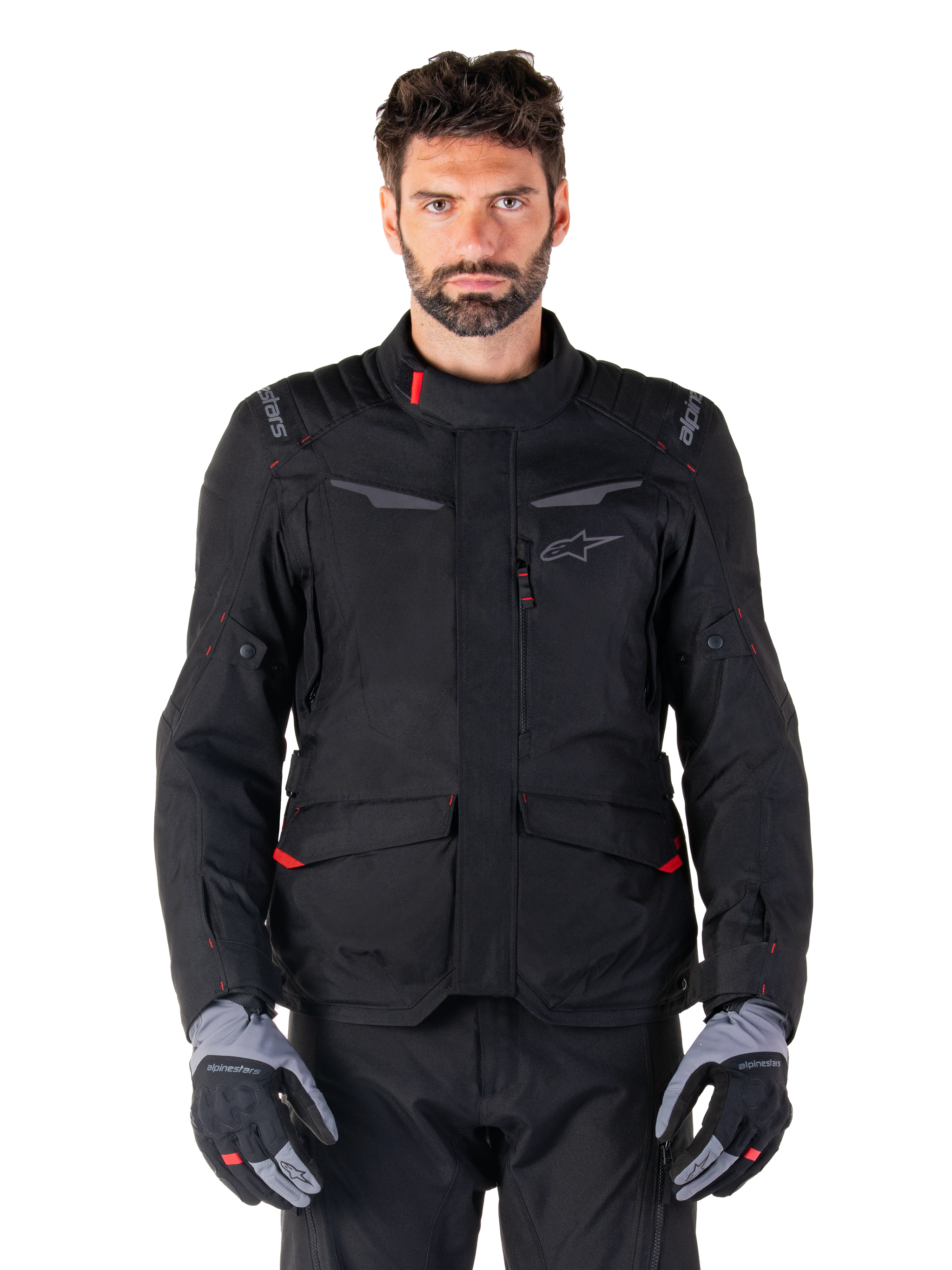 Veste imperméable Alpinestars ST-1, veste de touring d'aventure moto noire, portée par un mannequin homme avec un pantalon noir assorti et des gants gris/noir, style minimaliste, logo Alpinestars sur les épaules, grandes poches cargo avec détails rouges, conception ergonomique pour le touring routier par tous les temps