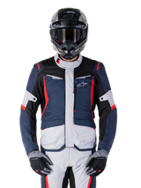 Veste imperméable Alpinestars ST-1, veste de tourisme d'aventure, bleu foncé, noir et rouge vif, portée avec un casque d'aventure noir et des gants gris, avec empiècements gris clair et accents rouges sur les épaules et les fermetures éclair, design épuré avec logo Alpinestars sur la poitrine, équipement de moto imperméable durable