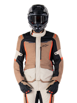Veste imperméable Alpinestars ST-1, Dark Khaki Sand Black Red Fluo, portée par un pilote avec un casque et des gants noirs, équipement de touring aventure avec empiècements beige et marron et accents rouge fluo vifs, vue de face sur fond blanc.