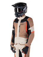 Veste imperméable Alpinestars ST-1, veste de touring aventure, kaki foncé, sable, noir et rouge fluo, portée par un motard avec un casque de motocross noir et des lunettes, dotée d'une conception multi-panneaux avec des poches cargo et le logo Alpinestars sur la poitrine