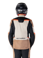 Veste imperméable Alpinestars ST-1, Dark Khaki Sand Black Red Fluo, vue arrière, veste de moto aventure touring dotée d'un large panneau dorsal couleur sable, de manches noires et d'accents rouge fluo, portée par un motard avec un casque noir.