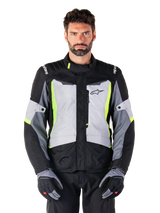 Veste imperméable Alpinestars ST-1, veste de moto adventure touring, Gris Glacé, Noir et Jaune Fluo, portée par un homme avec des gants assortis et un pantalon noir, dotée de détails jaune fluo sur la poitrine et le col, logo Alpinestars sur la poitrine