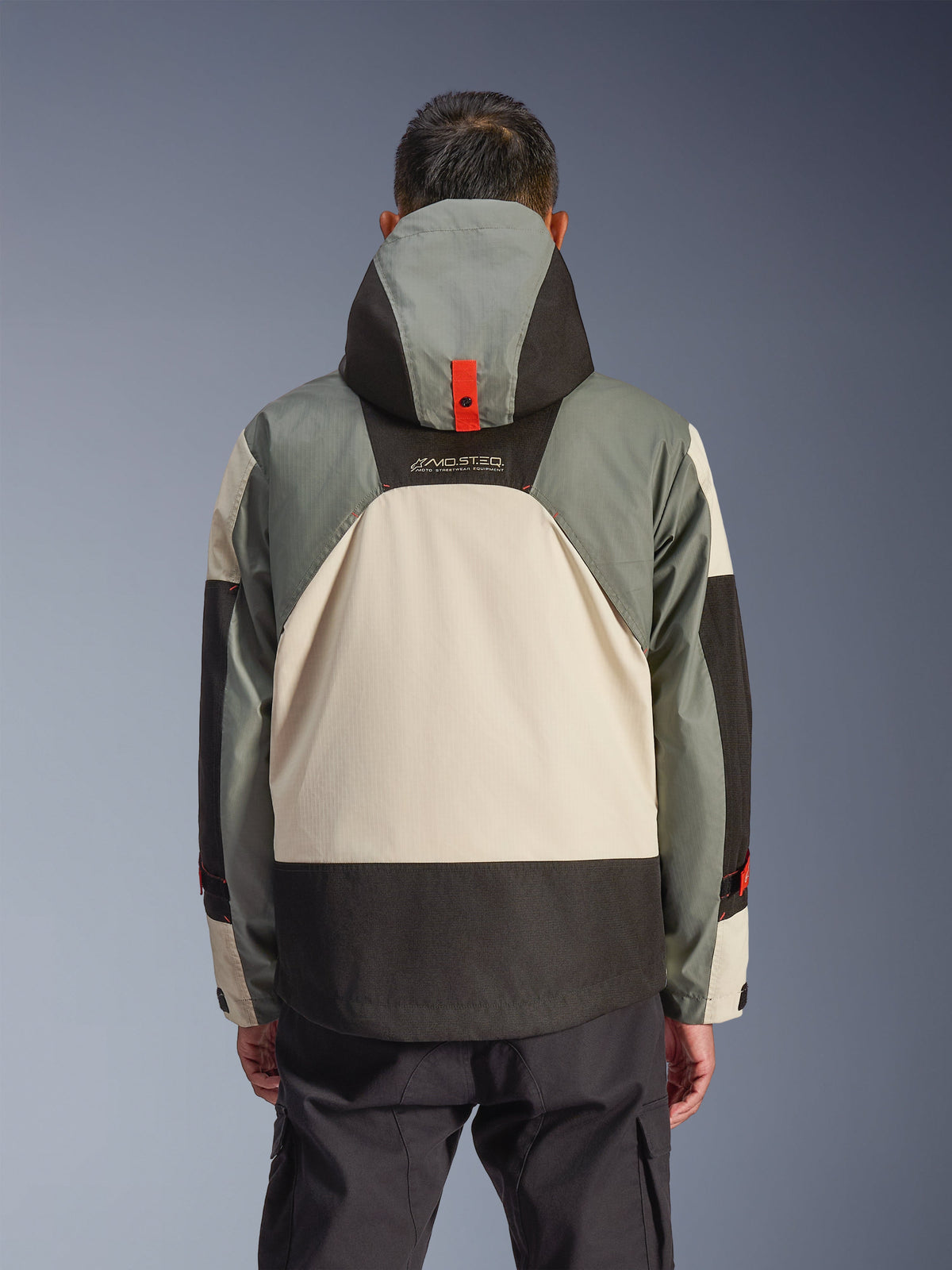 Vue arrière de la veste imperméable Alpinestars Mospher en Fur Gray Black Ice, présentant un design à empiècements gris, crème et noir, silhouette de parka à capuche avec détail d'ancrage à bouton-pression rouge, marquage « MO.ST.EQ. », associée à un pantalon de moto cargo noir