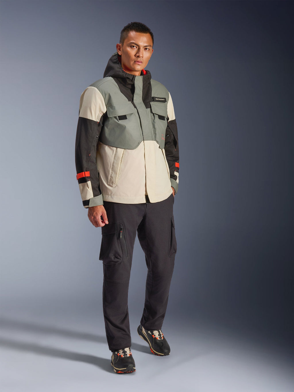 Veste imperméable Alpinestars Mospher en Fur Gray Black Ice, parka de moto technique multicolore avec panneaux gris, noirs et blanc cassé et touches de rouge, design fonctionnel avec grandes poches et capuche, portée avec un pantalon cargo gris foncé et des baskets noires, équipement de moto urbain contemporain