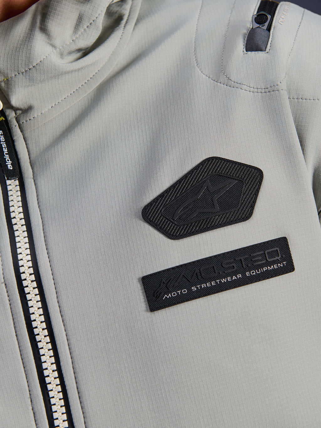 Alpinestars Moblast Waterproof Jacket, Iron Gray, close-up detail of chest patches featuring the Alpinestars logo and MO.ST.EQ. Moto Streetwear Equipment branding, technical ripstop fabric with heavy-duty front zipper and shoulder pocket detail.