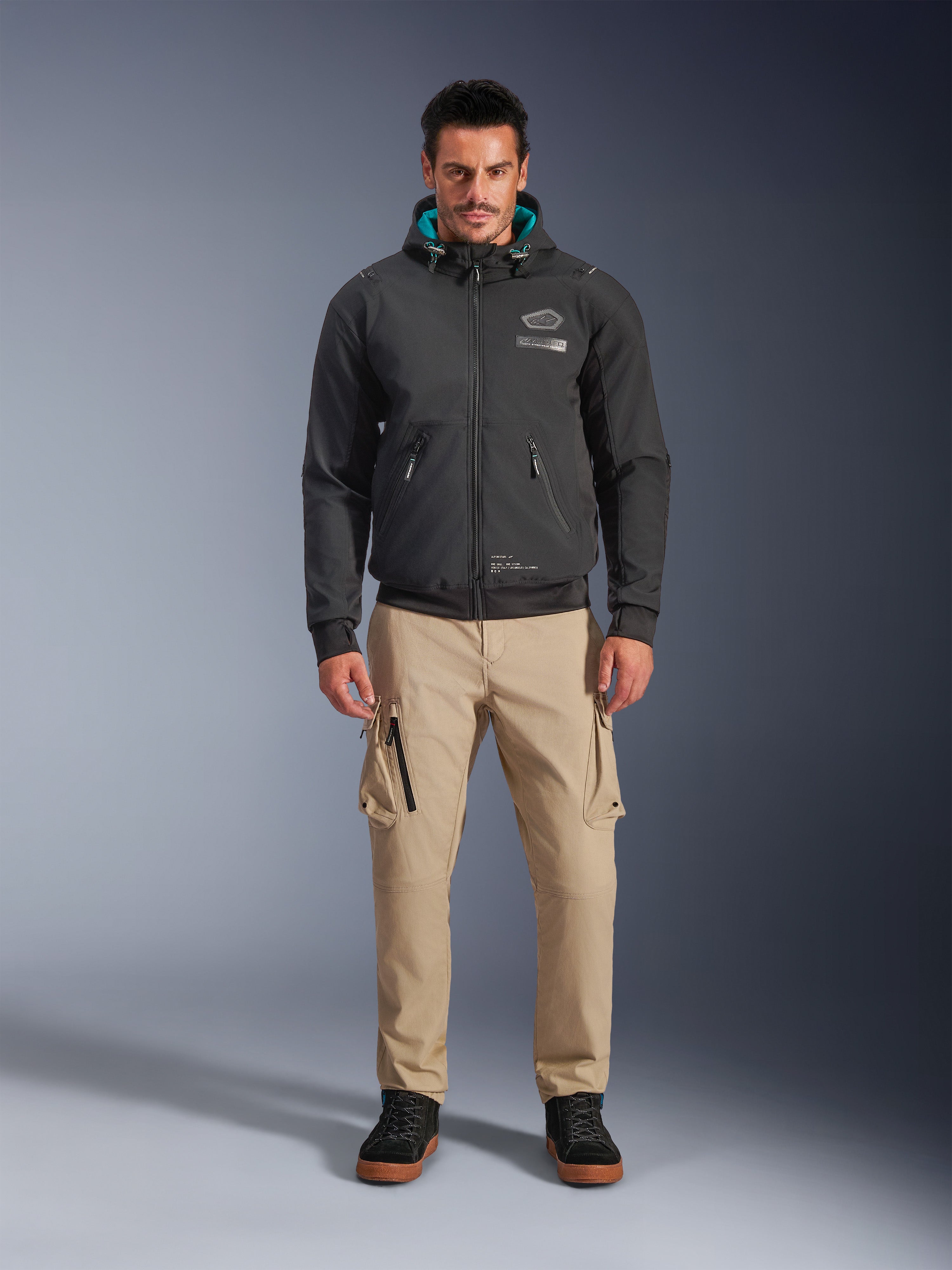 Veste imperméable Alpinestars Moblast, hoodie de moto urbain noir avec doublure de capuche bleu sarcelle, porté avec un pantalon cargo beige et des bottes montantes noires, vêtement de moto contemporain toutes saisons