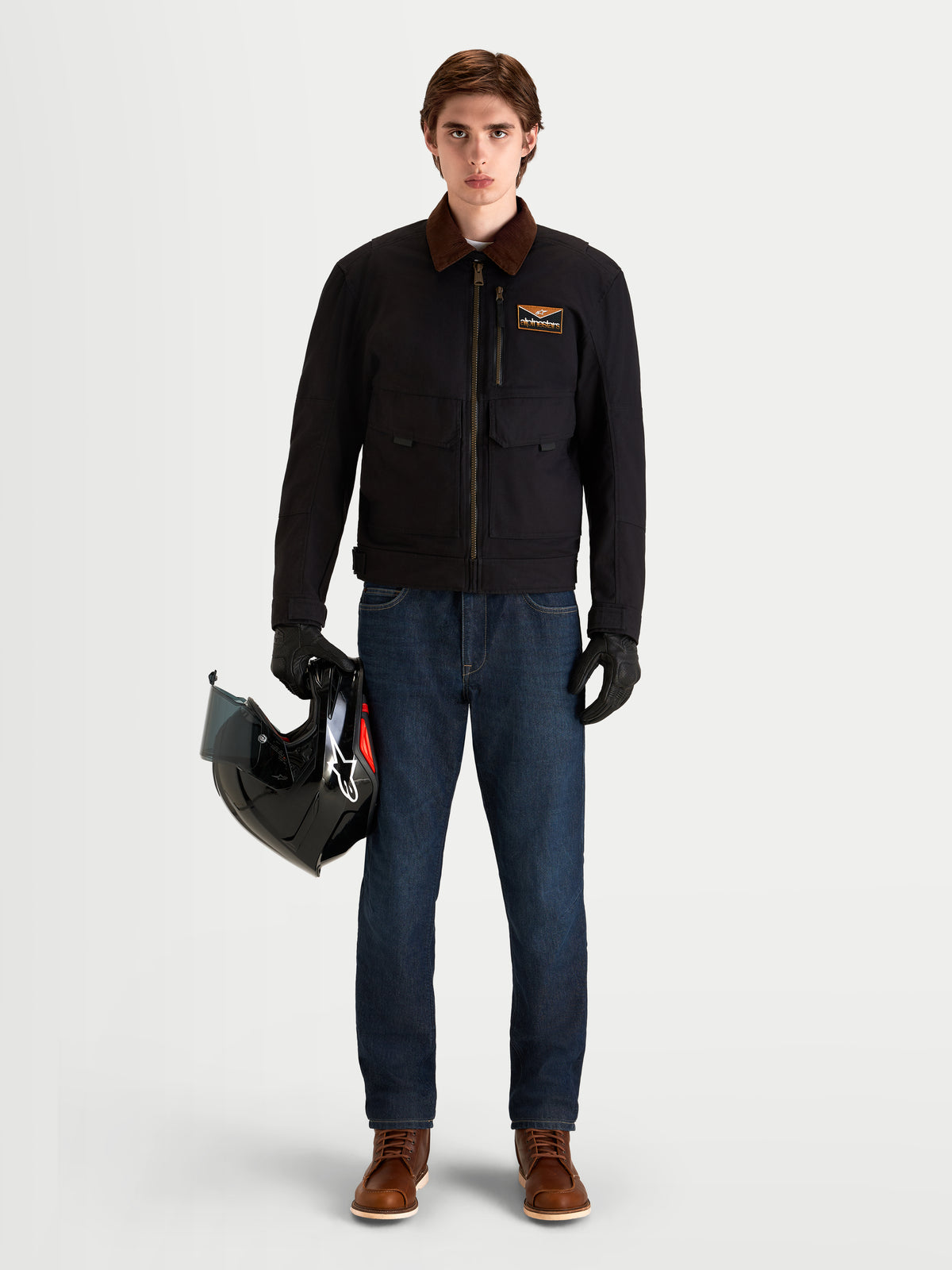 Alpinestars Mustang Waterproof Jacket, men's urban contemporary motorcycle jacket in dark anthracite and dark brown, front view on a model wearing protective riding gear, holding a helmet, showcasing workwear-inspired stretch twill construction and corduroy collar