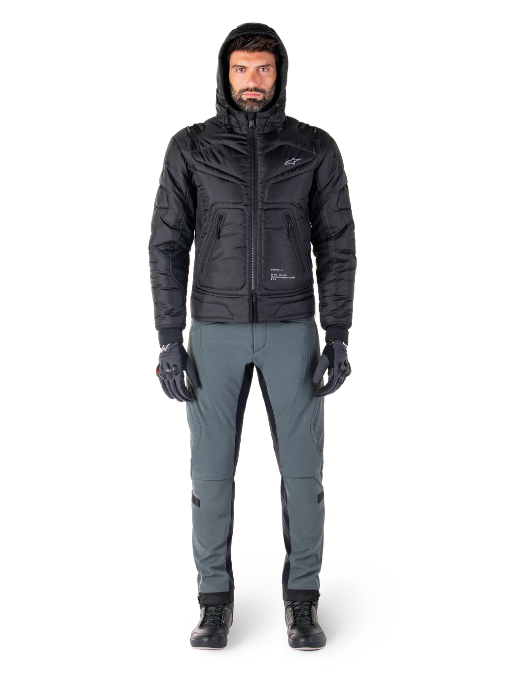 Veste Alpinestars Mohobbs Waterproof noire, mannequin portant un blouson aviateur matelassé à capuche, associé à un pantalon de moto gris, des gants noirs et des bottes noires, équipement de moto urbain contemporain