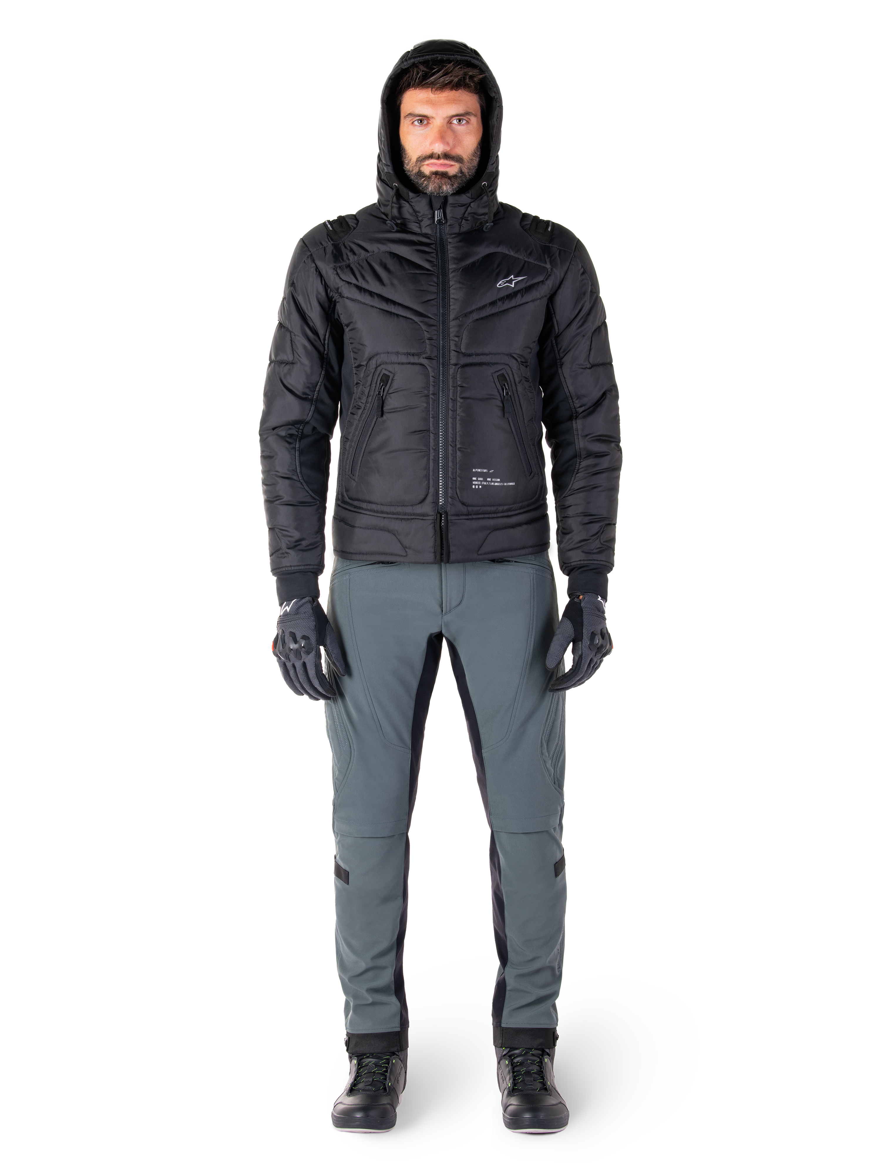 Veste Alpinestars Mohobbs Waterproof noire, mannequin portant un blouson aviateur matelassé à capuche, associé à un pantalon de moto gris, des gants noirs et des bottes noires, équipement de moto urbain contemporain