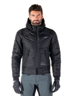 Veste Alpinestars Mohobbs Waterproof, blouson aviateur de moto rembourré noir, tissu poly matelassé avec fermeture éclair centrale et poches zippées, petit logo blanc sur la poitrine, porté par un mannequin avec un pantalon gris et des gants, design streetwear urbain pour la conduite en ville.