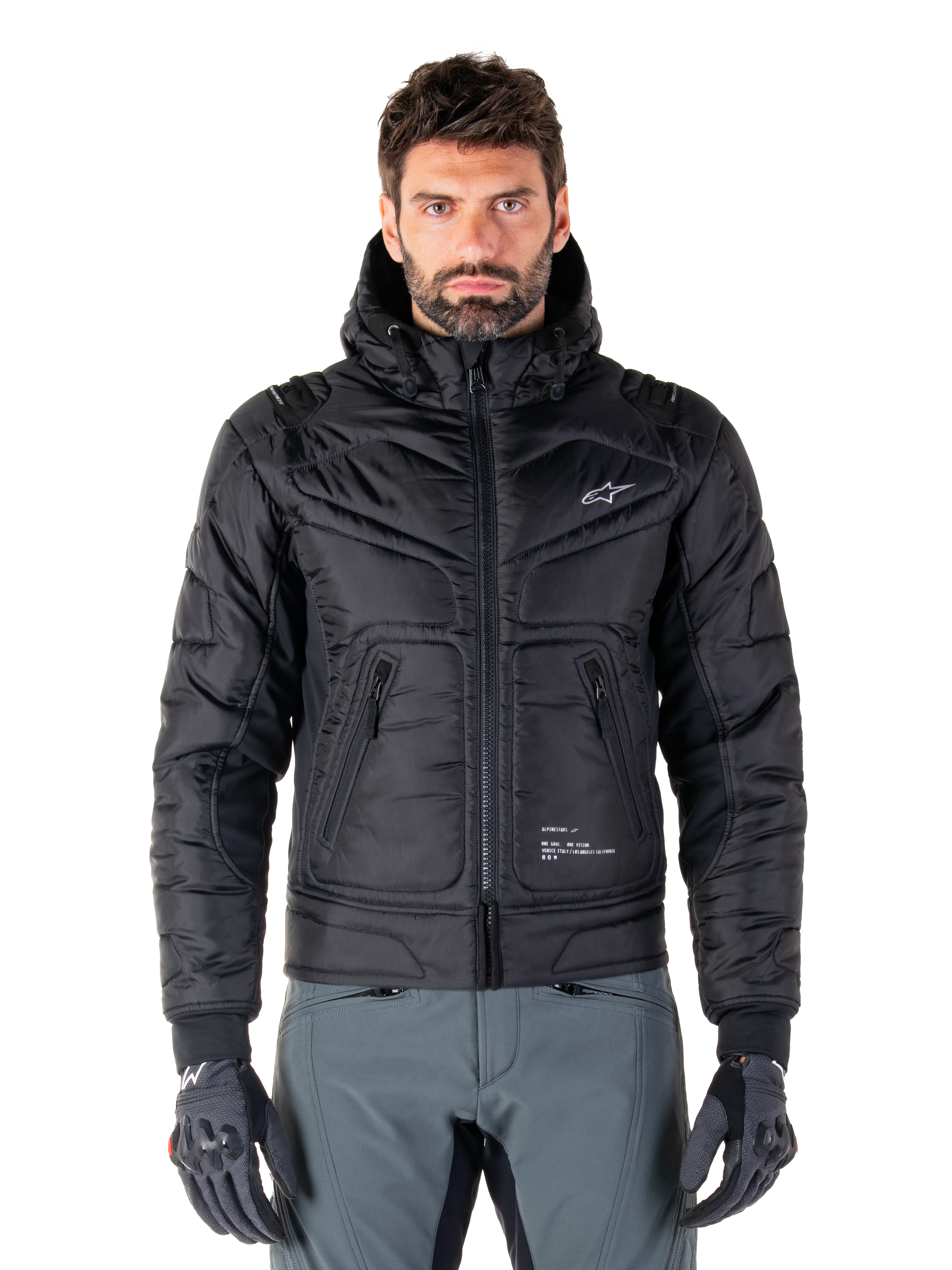 Veste Alpinestars Mohobbs Waterproof, blouson aviateur de moto rembourré noir, tissu poly matelassé avec fermeture éclair centrale et poches zippées, petit logo blanc sur la poitrine, porté par un mannequin avec un pantalon gris et des gants, design streetwear urbain pour la conduite en ville.