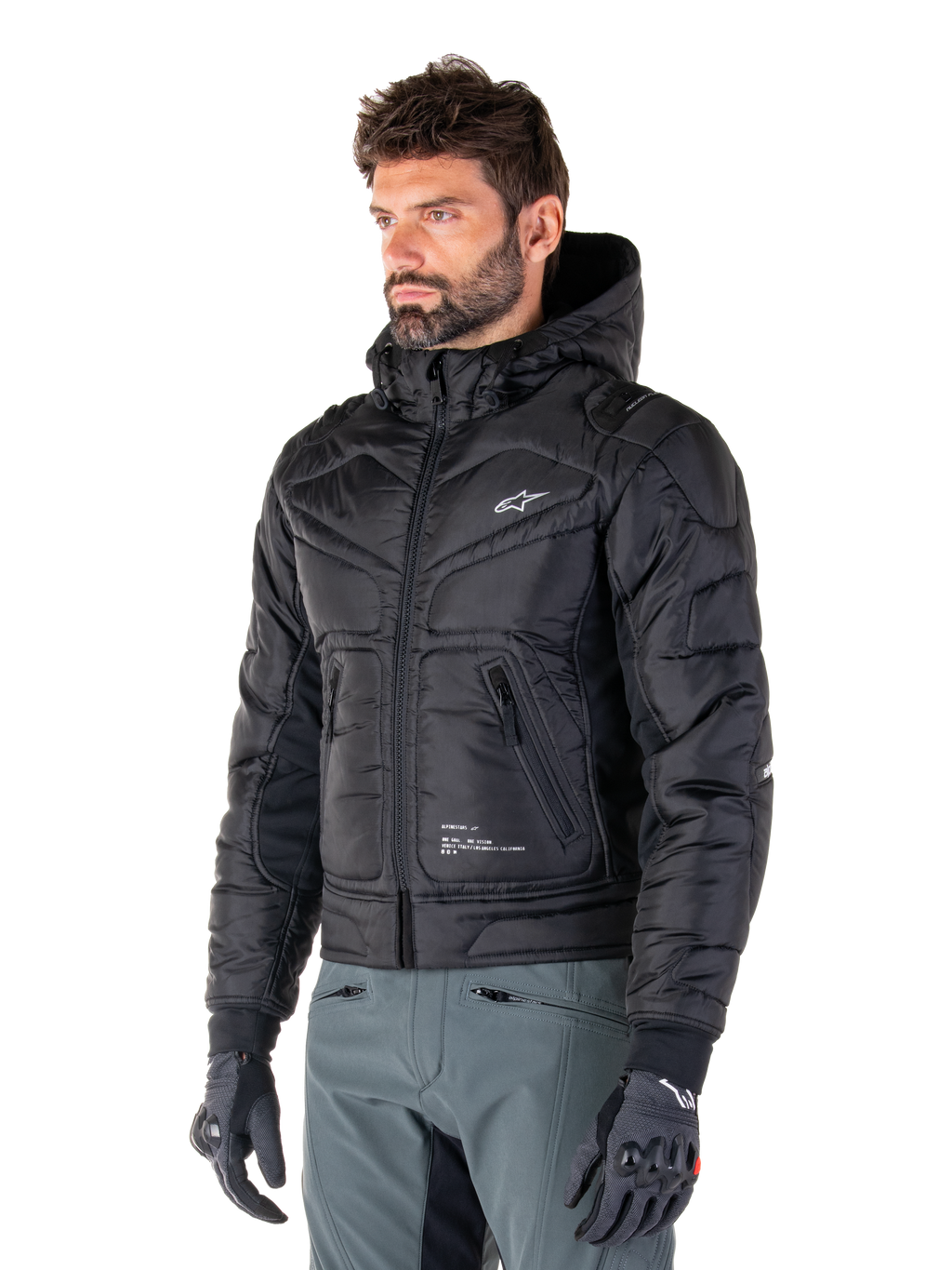 Veste imperméable Alpinestars Mohobbs, noire, design bomber matelassé et rembourré avec une capuche, portée par un homme avec un pantalon de moto gris et des gants noirs, vêtement de moto d'inspiration streetwear urbain arborant un petit logo blanc sur la poitrine.