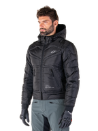 Veste imperméable Alpinestars Mohobbs, noire, design bomber matelassé et rembourré avec une capuche, portée par un homme avec un pantalon de moto gris et des gants noirs, vêtement de moto d'inspiration streetwear urbain arborant un petit logo blanc sur la poitrine.