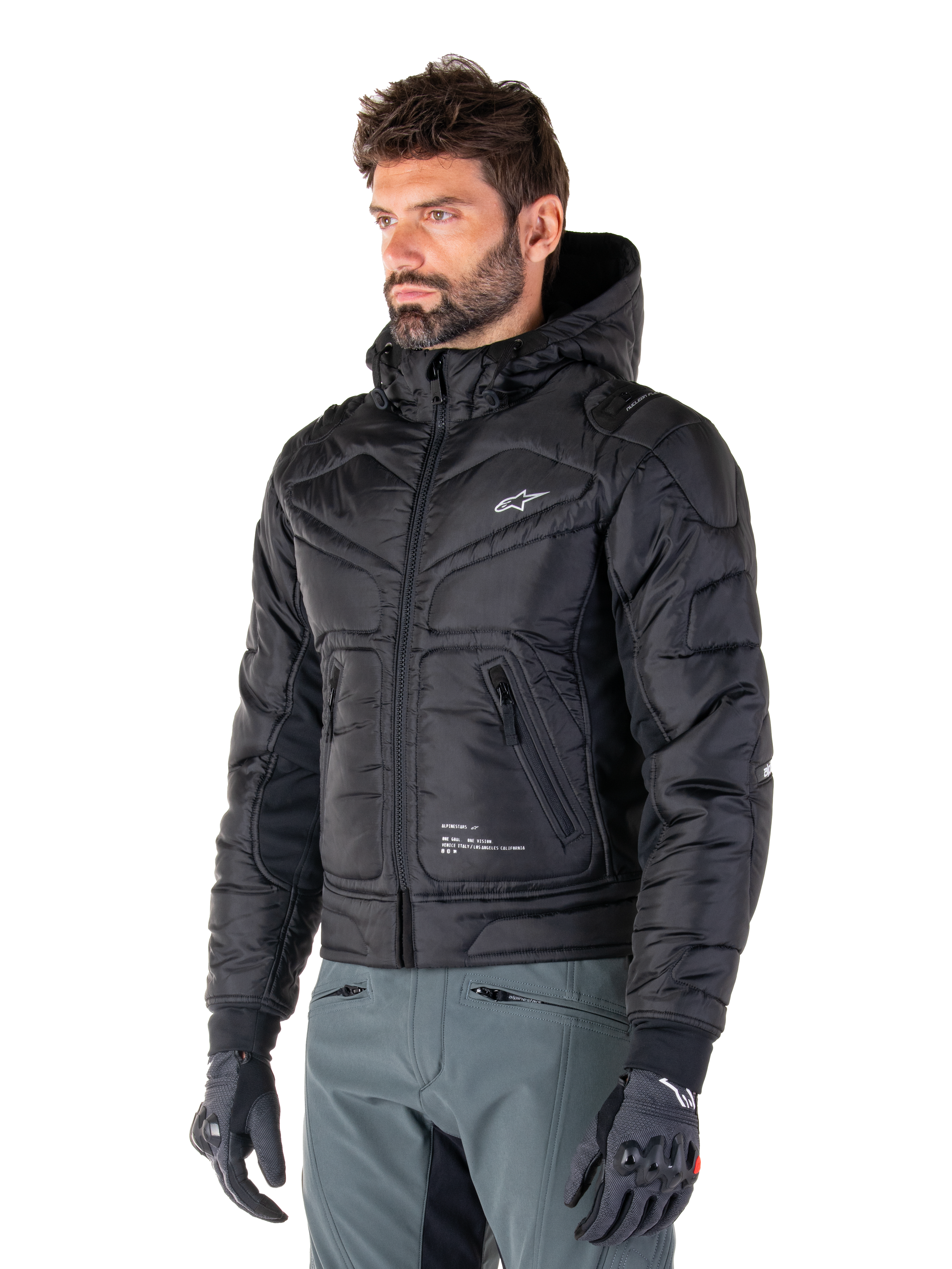 Veste imperméable Alpinestars Mohobbs, noire, design bomber matelassé et rembourré avec une capuche, portée par un homme avec un pantalon de moto gris et des gants noirs, vêtement de moto d'inspiration streetwear urbain arborant un petit logo blanc sur la poitrine.