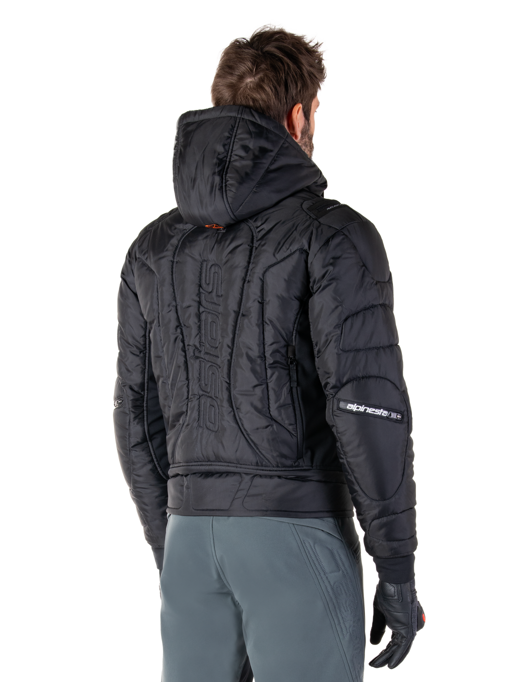 Veste imperméable Alpinestars Mohobbs, noire, vue arrière montrant la construction matelassée et rembourrée, une capuche, le mot-symbole astars vertical au dos et le marquage Alpinestars sur les manches, portée avec un pantalon gris et des gants noirs.