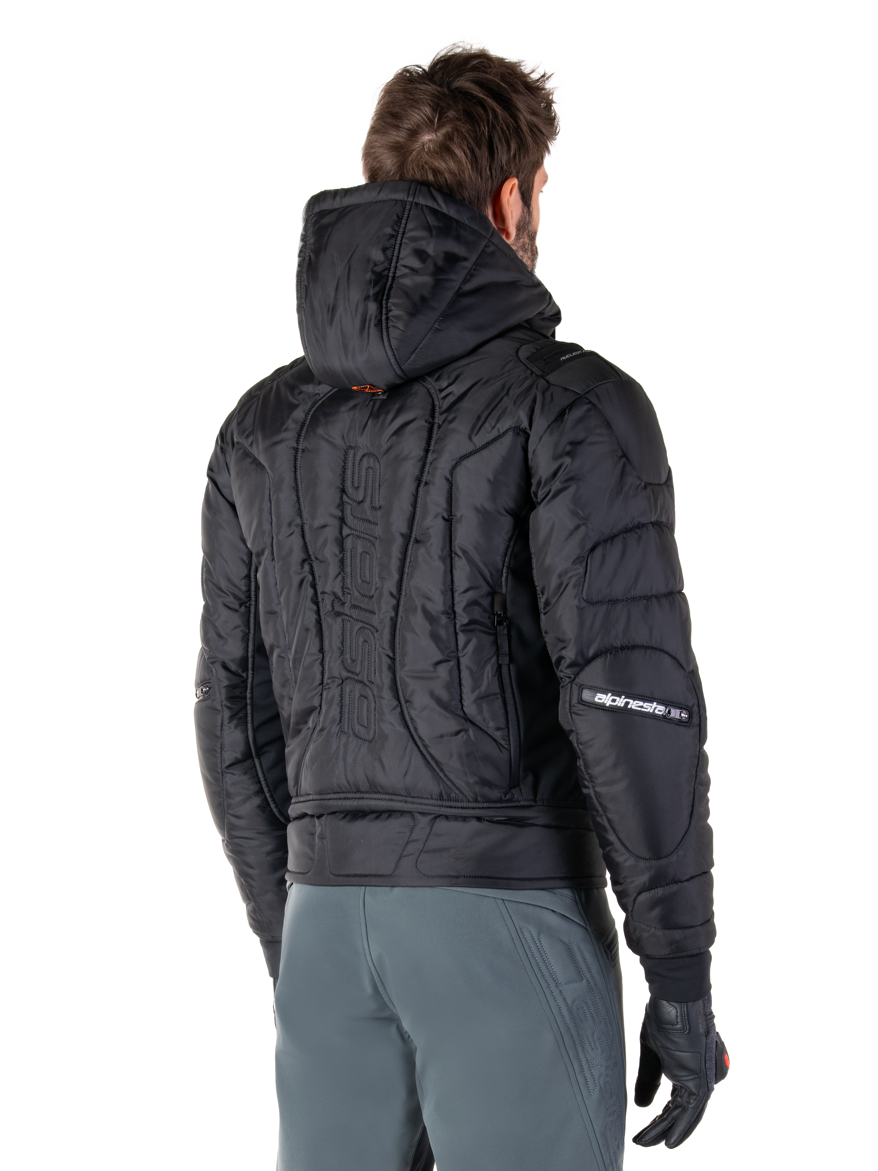 Veste imperméable Alpinestars Mohobbs, noire, vue arrière montrant la construction matelassée et rembourrée, une capuche, le mot-symbole astars vertical au dos et le marquage Alpinestars sur les manches, portée avec un pantalon gris et des gants noirs.