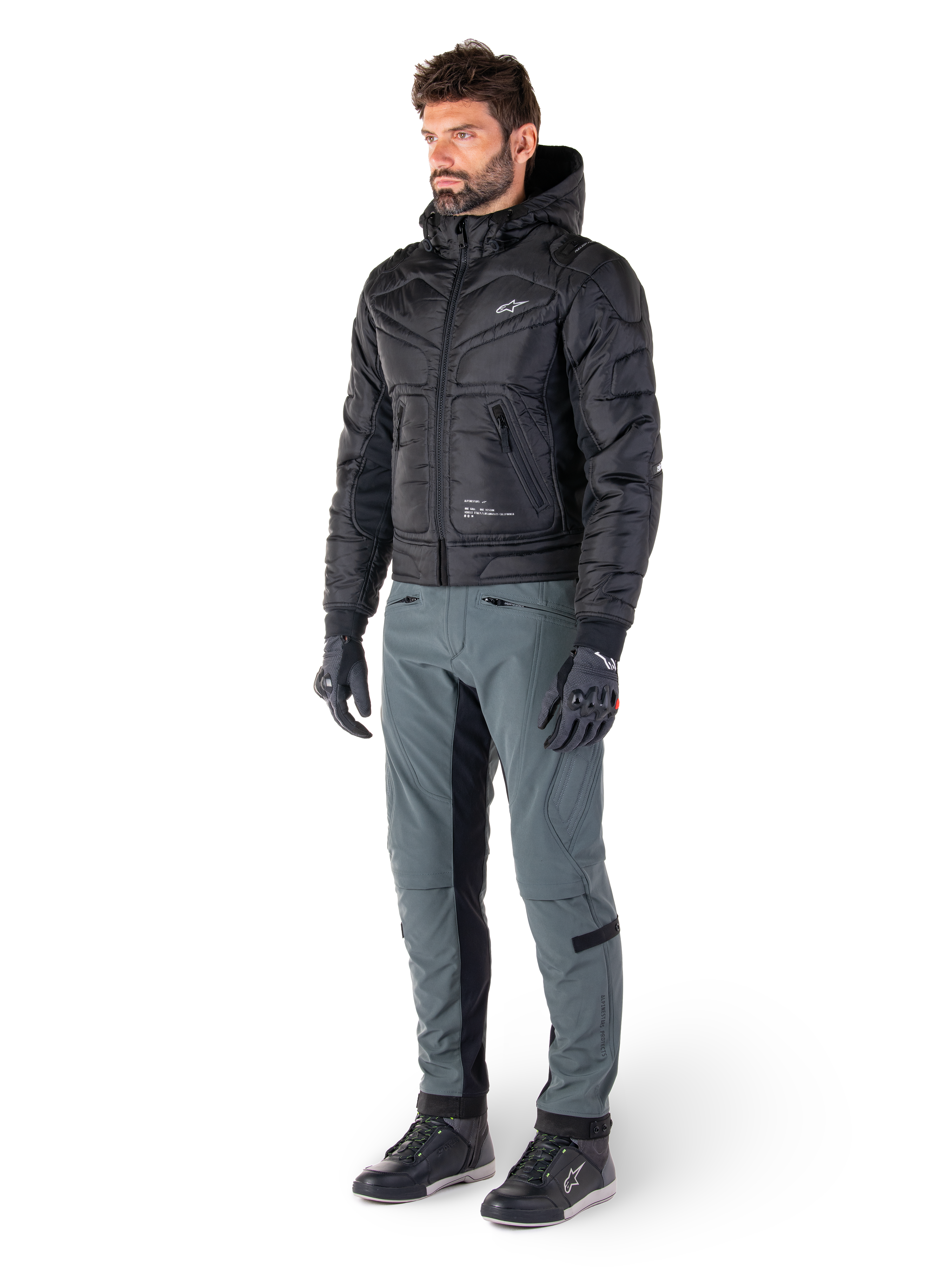 Veste Mohobbs Waterproof Alpinestars, noire, blouson aviateur de moto urbain matelassé avec capuche, portée par un mannequin avec un pantalon de moto gris et des gants noirs, avec un logo blanc sur la poitrine, équipement de protection moderne d'inspiration streetwear
