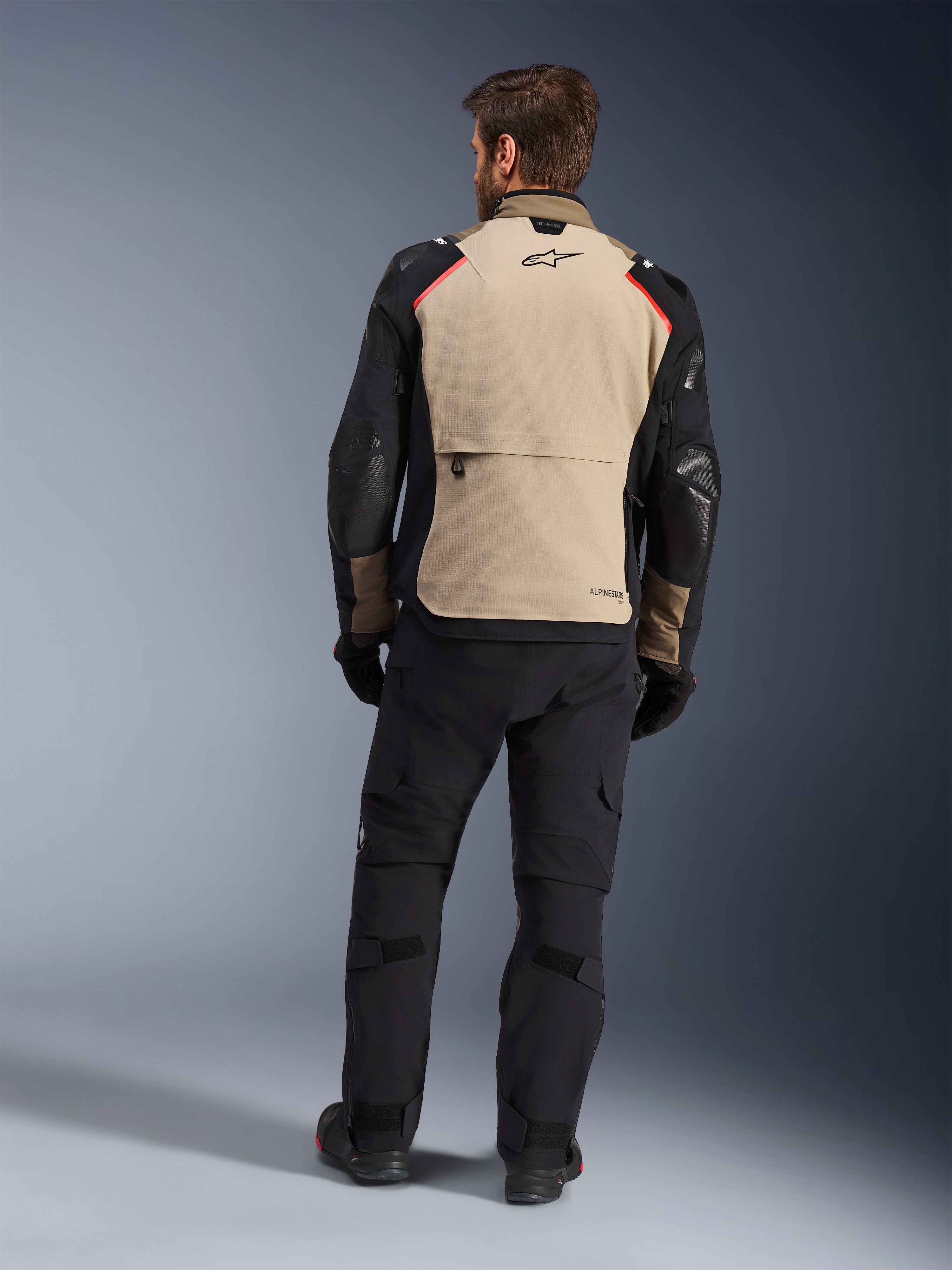 Veste Alpinestars Andes Pro Drystar® XF, Walnut Aluminum, vue de dos, veste de moto d'aventure avec manches noires et accents rouges aux épaules, portée avec un pantalon de moto et des gants noirs.