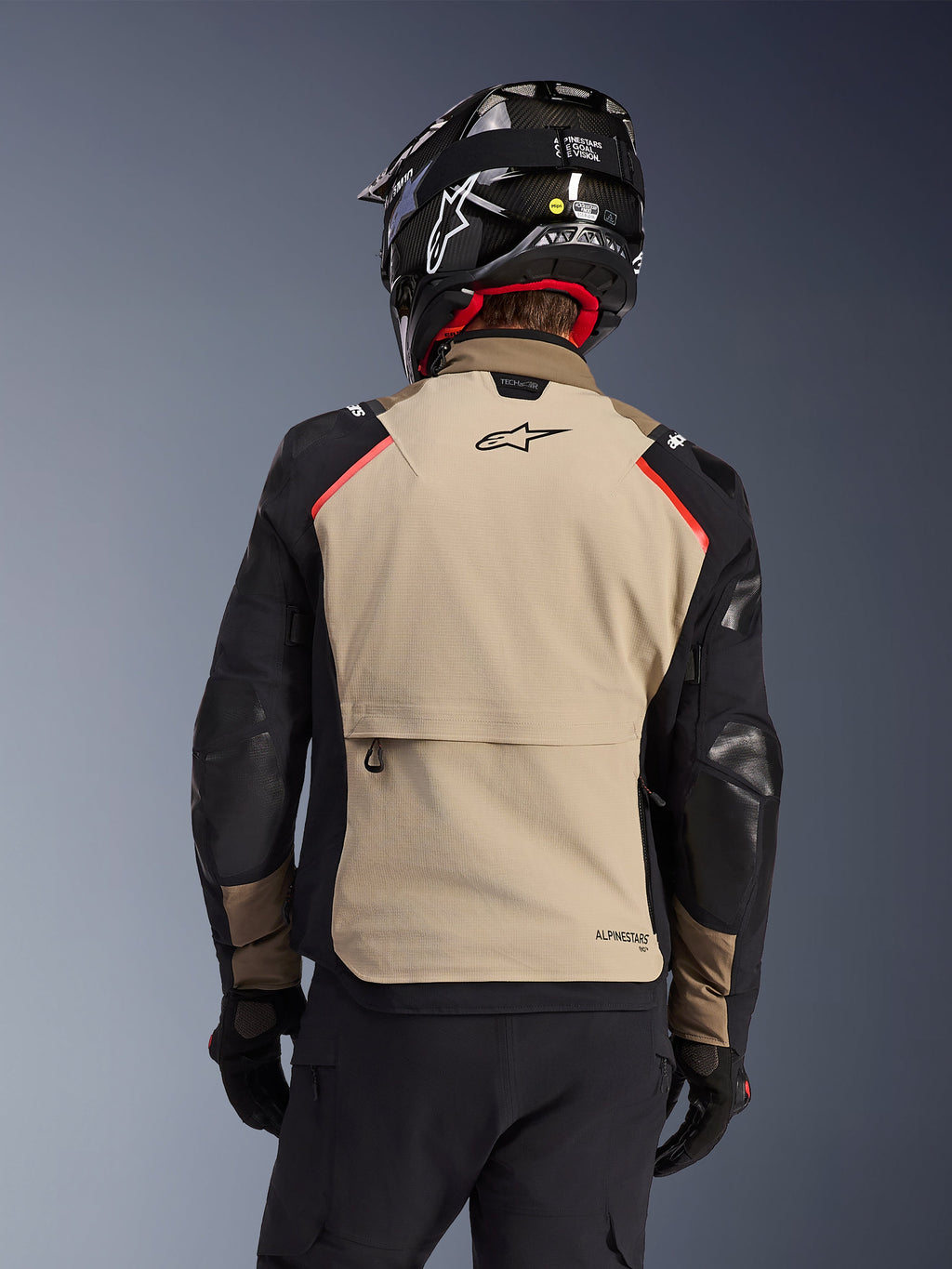 Veste Alpinestars Andes Pro Drystar® XF en Walnut Aluminum, vue de dos, panneau arrière beige avec grande poche cargo, manches noires avec empiècements renforcés, logo Alpinestars sur le haut du dos, associée à un casque et des gants noirs pour le touring d'aventure.