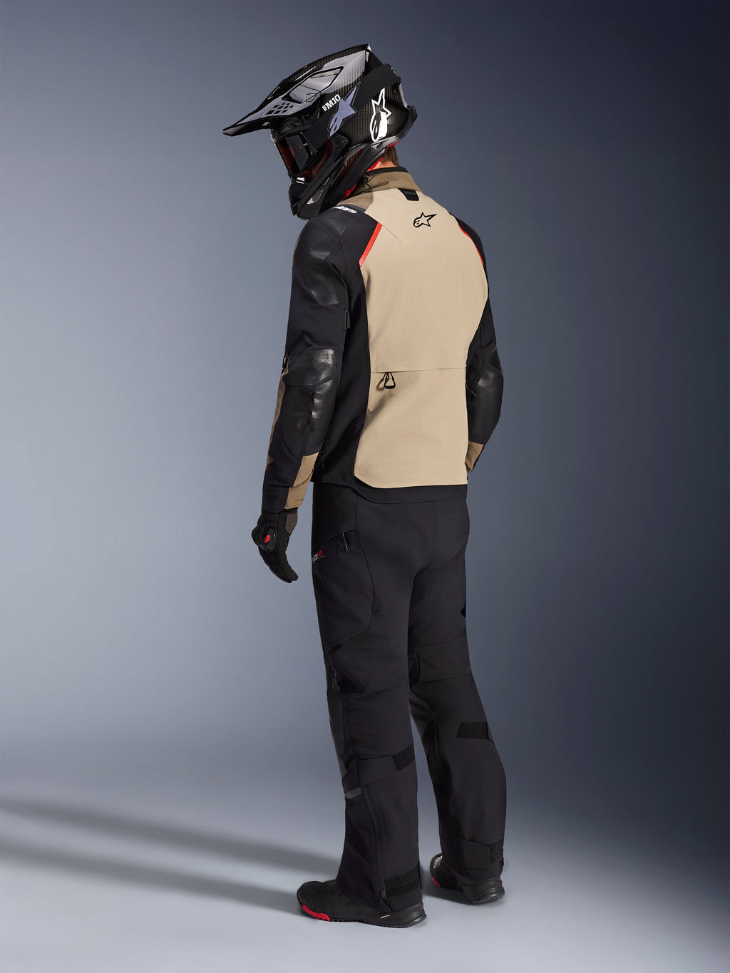 Motard vu de dos portant la veste Alpinestars Andes Pro Drystar® XF en coloris Walnut Aluminum, avec un panneau arrière beige, des manches noires et des touches de rouge, casque d'aventure noir, pantalon de moto noir et bottes noires, équipement professionnel d'aventure touring.
