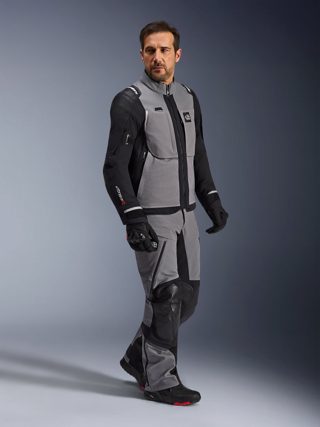 Veste Alpinestars Andes Pro Drystar® XF, veste de moto d'aventure, gris foncé et noir, portée par un mannequin avec un pantalon en textile assorti et des gants noirs, dotée d'une conception multi-poches, d'une coque laminée technique à 3 couches et d'une coupe articulée pour les longs trajets d'aventure.