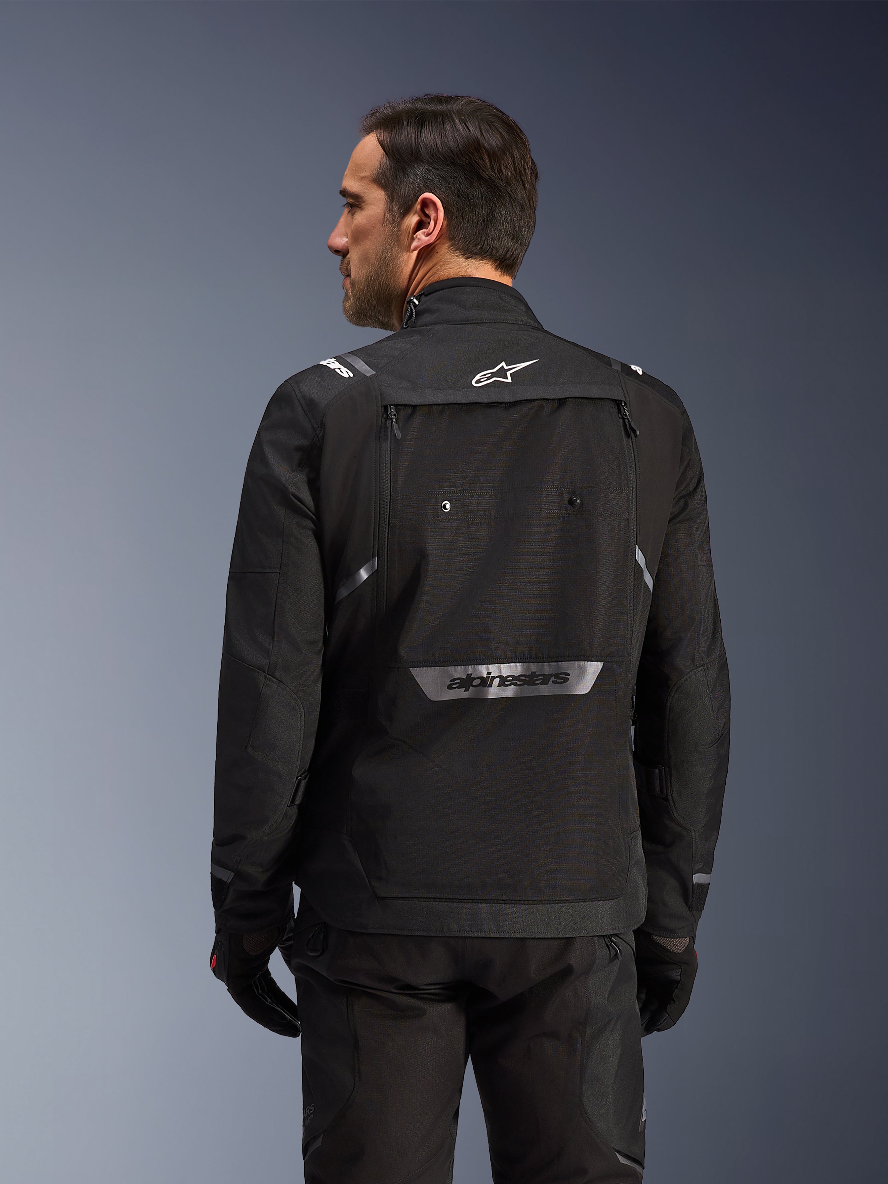 Rider from behind wearing Alpinestars Cusco Drystar® Jacket in Black, textile adventure touring jacket with large rear storage pocket and reflective branding, paired with matching black pants and gloves