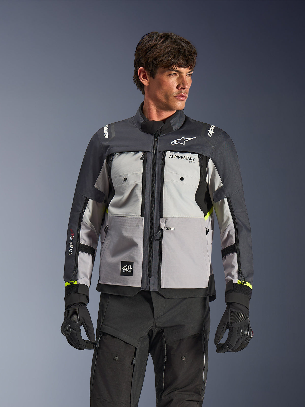 Veste Alpinestars Cusco Drystar®, veste de moto adventure touring, gris glace, gris moyen et gris foncé, portée par un mannequin avec un pantalon et des gants noirs, présentant un design textile technique multi-panneaux, des poches de poitrine, le marquage « Drystar » sur la manche et des touches discrètes de jaune fluo aux poignets.