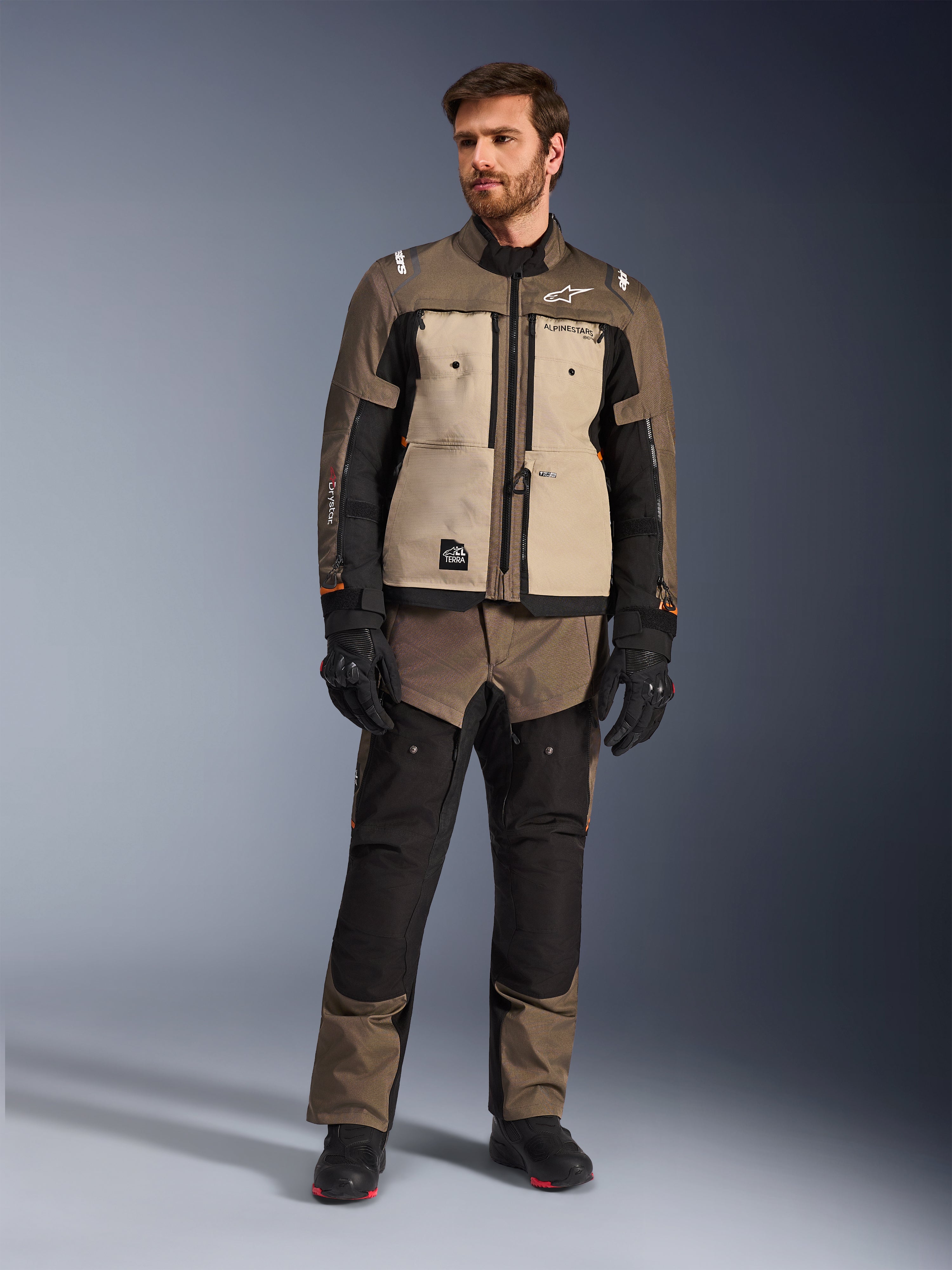 Alpinestars Cusco Drystar® Jacket in Walnut Aluminum Black, worn by a male rider with matching adventure touring pants, black gloves, and boots, featuring tan chest panels, black sleeves with subtle orange accents, and functional ventilation zippers for motorcycle road riding.