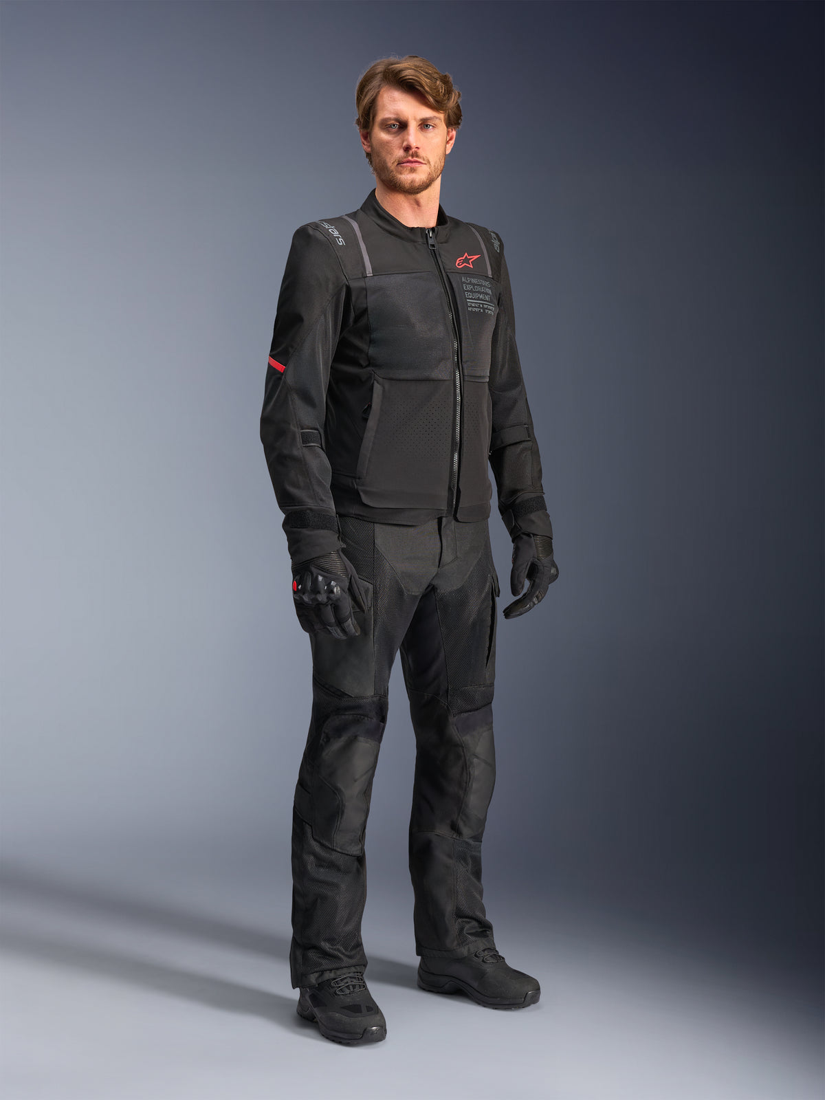 Modèle masculin portant la veste Alpinestars ST-2 Air noire, dotée d'un logo étoile rouge et du marquage 'Alpinestars Exploration Equipment', associée à un pantalon de moto, des gants et des bottes noirs assortis pour une tenue de touring estivale moderne.