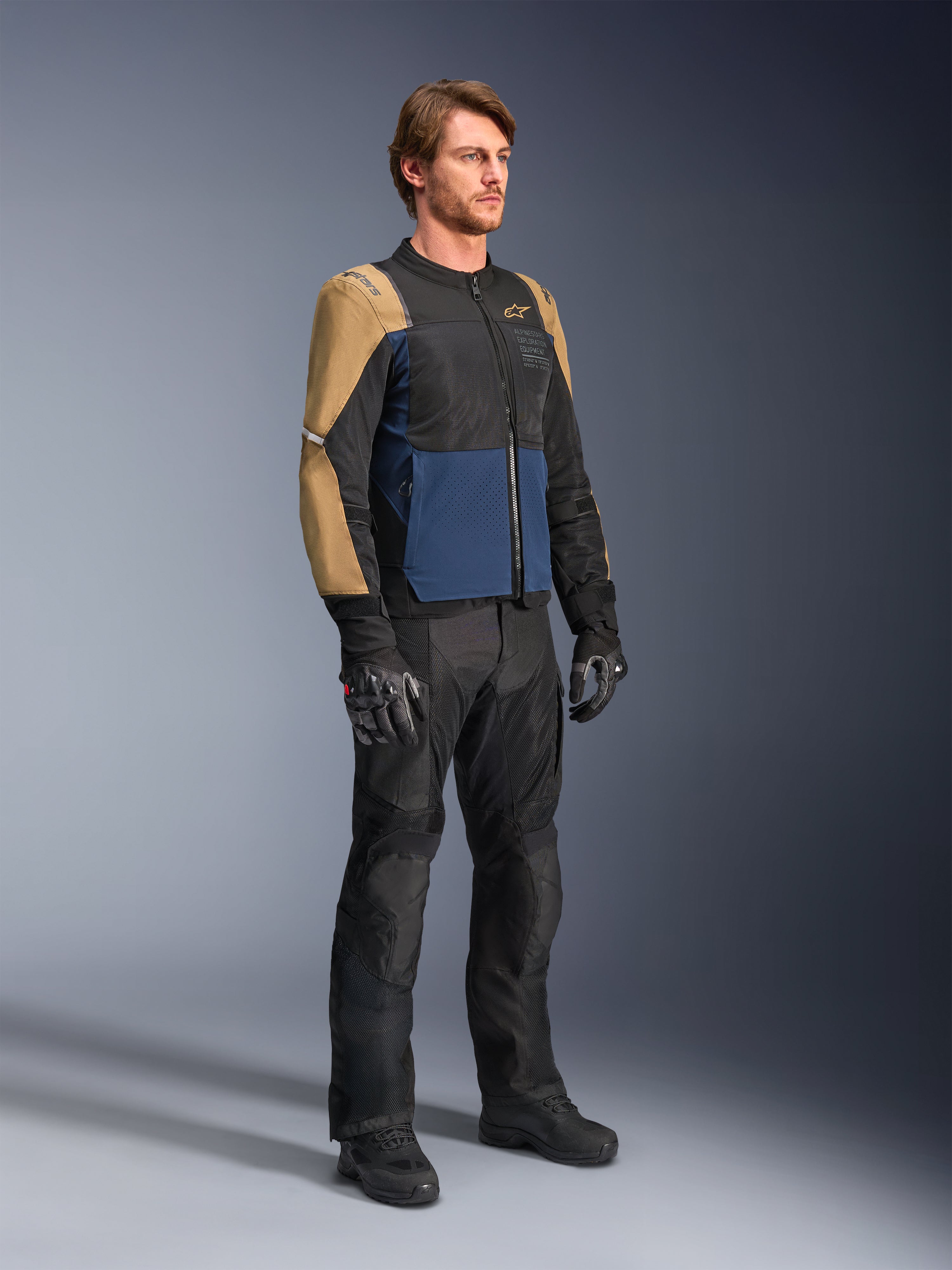 Motard portant la veste Alpinestars ST-2 Air en bleu profond, kaki foncé et noir, dotée de larges panneaux en mesh et de perforations au laser, associée à un pantalon, des gants et des bottes de moto noirs pour un look de tourisme d'été complet.