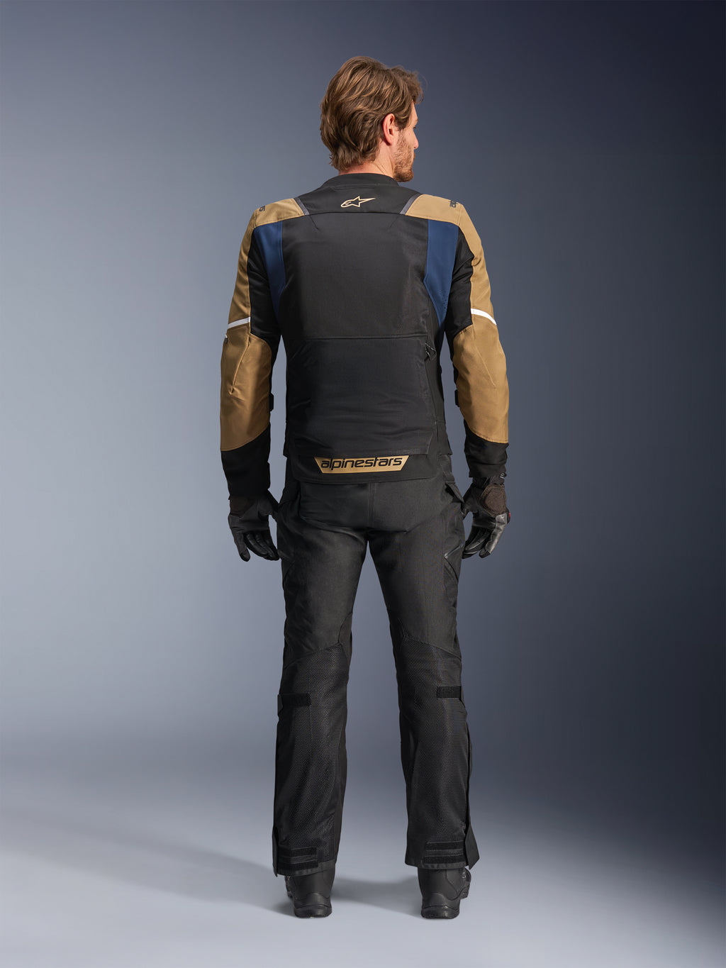 Veste Alpinestars ST-2 Air en bleu profond, kaki foncé et noir, vue arrière sur modèle montrant le large panneau dorsal en mesh et les manches kaki avec rayures blanches, associée à un pantalon technique et des gants noirs pour le touring d'aventure.